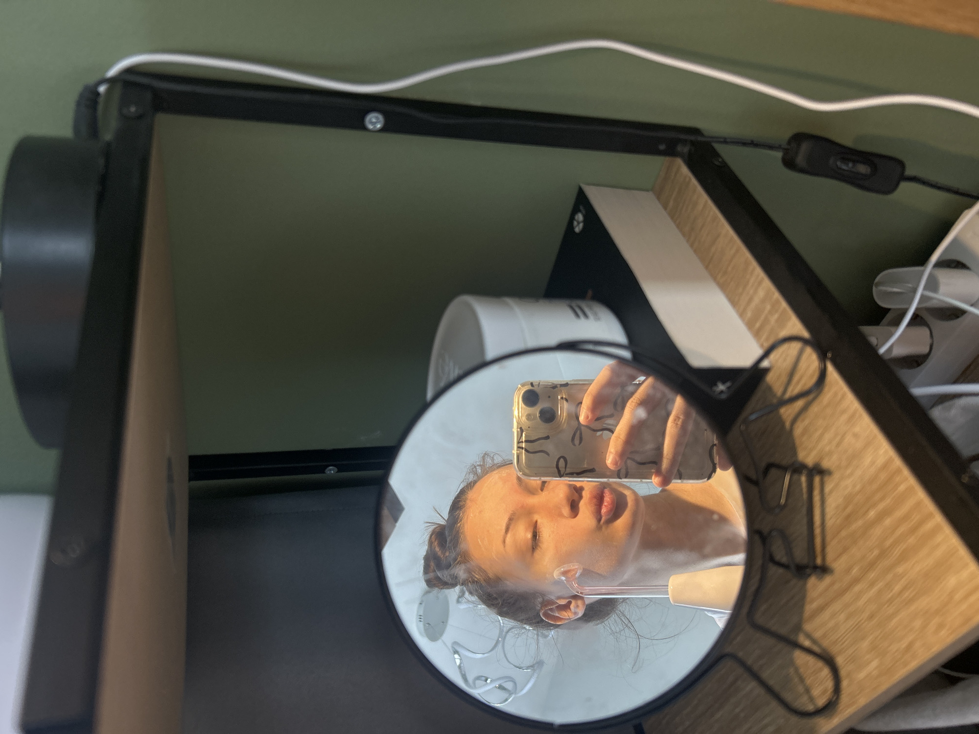 A mirror reflecting a woman taking a selfie is the central element of the image. The woman, visible in the mirror's reflection, holds a phone with a patterned case. Part of her face is obscured by the phone, but her closed eyes, nose, and lips are visible. She is using an electrical facial massager and has her hair in a bun.
The mirror itself has a black, round frame and a wire stand. The background contains parts of furniture, including wood-grained surfaces, a dark-colored shelf, and a white electrical cord.
The image does not offer any information or landmarks that would suggest it was taken in Burgas, Bulgaria.