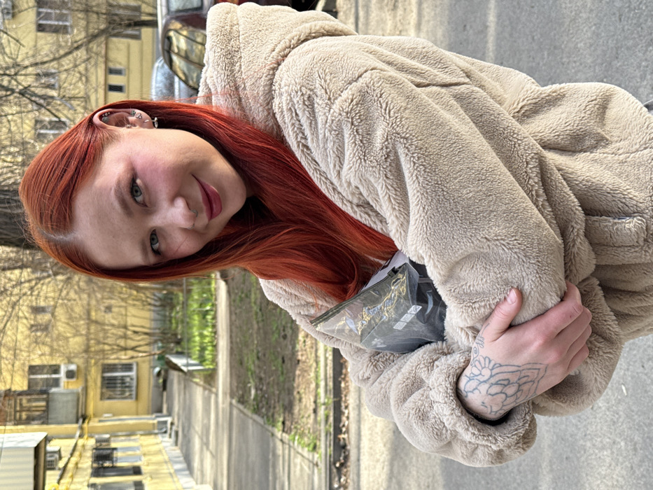 A young adult female with red hair, fair complexion, and prominent pink blush is depicted standing outdoors in Rostov-on-Don, Russia. She wears a light beige, hooded, faux-shearling jacket, her arms crossed, holding a dark, translucent plastic package containing unidentifiable geometric items against her chest. A light-colored tattoo is visible on her left hand. Her left ear displays multiple silver piercings, and a silver stud is present in her left nostril. She has green/blue eyes and red lipstick, looking directly at the camera with a slight smile. The background features a multi-story yellow residential building with multiple windows and balconies on the left, and bare deciduous trees. A paved sidewalk runs along the left side, with patches of green grass visible beneath the trees. A dark-colored vehicle is partially visible parked in the distant right background. The scene is illuminated by daylight, casting distinct shadows.