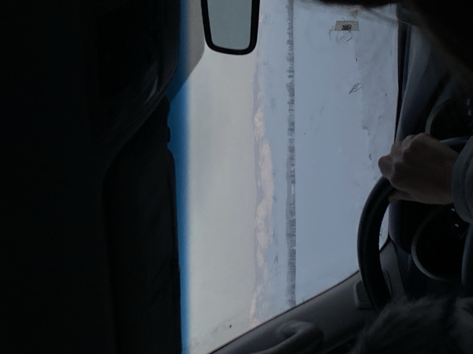 A view from inside a vehicle shows a driver with their hand on the steering wheel. The windshield is somewhat obscured, but a mountain range or hill is visible in the distance. A rearview mirror hangs from above. The car's interior is dark, contrasting with the brighter outdoor view. The scene appears to be a wintery or overcast day.