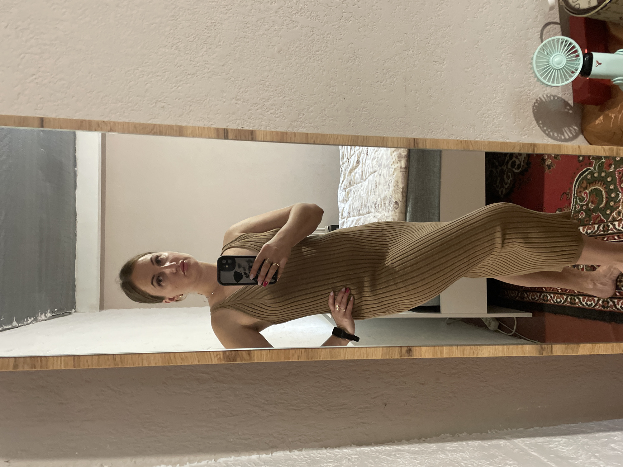 The image shows a woman standing in front of a full-length mirror in what appears to be a bedroom. The woman is wearing a brown, ribbed, sleeveless midi dress. She has her hair pulled back and is holding a smartphone in her right hand, taking a selfie. Her left hand is on her hip. In the reflection of the mirror, a bed with a patterned duvet is visible, along with a rug on the floor. To the right of the mirror, a small, light blue fan is on a surface. The room has light-colored walls and a textured ceiling. The image does not provide any specific indicators of the city of Zarichne, Ukraine.