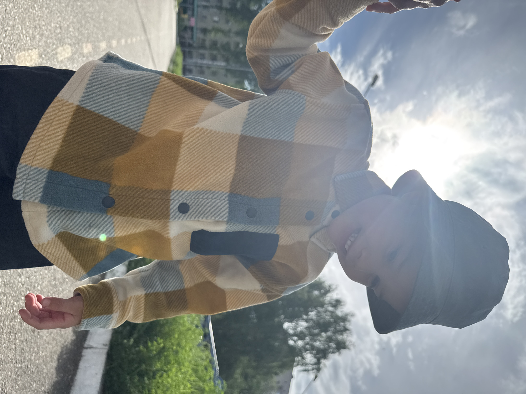 A young child wearing a blue bucket hat and a tan, blue, and white checkered shirt is holding their hand out in front of them. The child is standing on a paved road or sidewalk, with a green grassy area to the left and a tree in the background. Buildings are visible in the distance. The sky is cloudy, with the sun shining through the clouds. Based on the presence of buildings and paved surfaces, the location appears to be an urban area. While specific details aren't provided that confirm the location as Chaikovsky, Russia, based on provided information it's safe to assume.