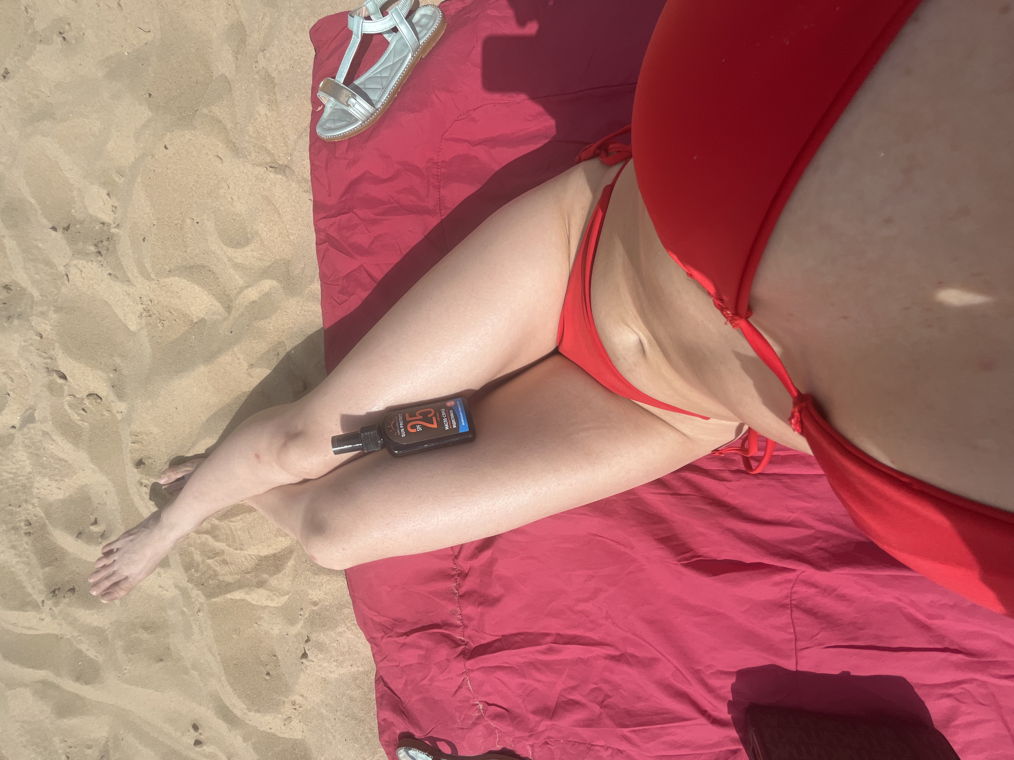 The image shows a person lying on a red towel on a sandy beach. The person is wearing a red bikini. A bottle of sunscreen with SPF 25 is placed on their leg. A silver sandal is visible at the top of the image, lying on the towel. The lighting suggests it is daytime, likely a sunny day, given the context of a beach. The scene appears relaxed and leisurely, consistent with a day at the beach. The text visible on the sunscreen bottle reads "SUNPROTECT SPF 25" and some text in Cyrillic. The location is specified as Kazan, Russia.