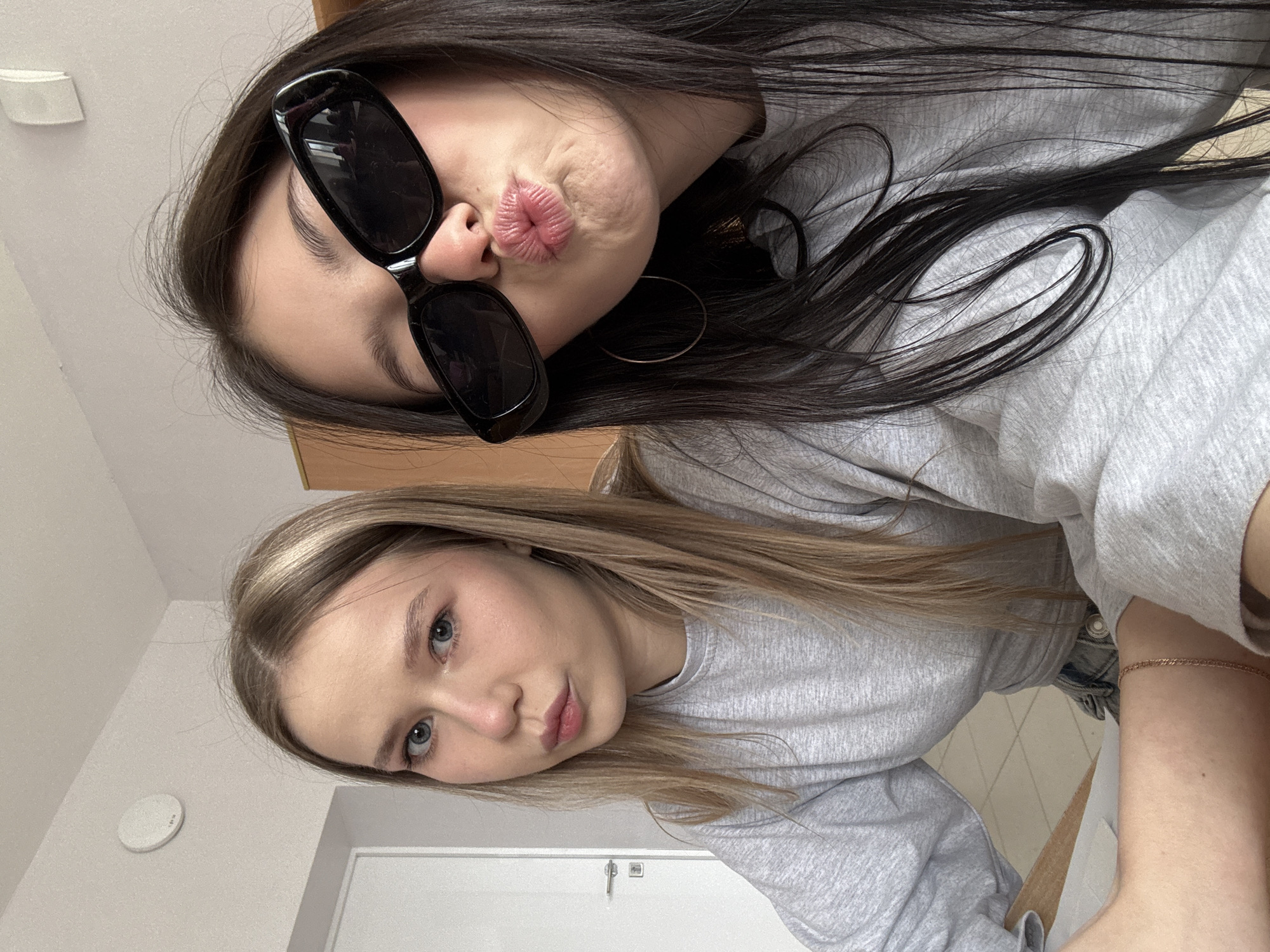 Two young women pose for a selfie in what appears to be an indoor setting. The woman at the top of the frame has dark hair and is wearing black sunglasses. She is making a kissing face. The woman at the bottom has blonde hair and blue eyes and is looking directly at the camera. Both women are wearing gray sweatshirts. The background includes a white ceiling with a smoke detector and a white door. While the city of Chaikovsky, Russia is mentioned, no identifiable features or signs in the image explicitly confirm this location. The setting appears to be a simple, possibly domestic, interior.