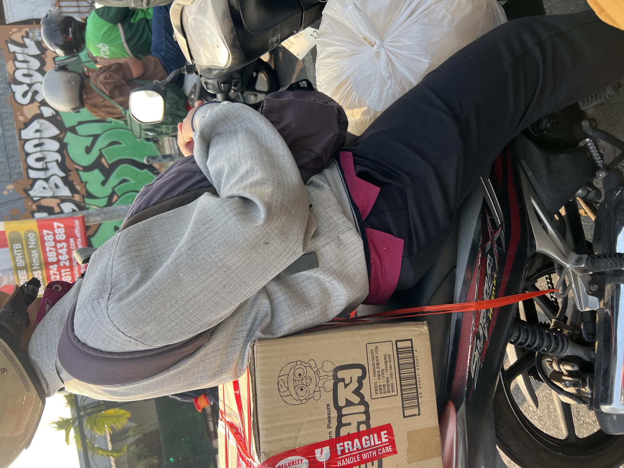 In the city of Sinduadi, Indonesia, a person wearing a helmet and a grey jacket is seated on a motorcycle, holding a cardboard box and a large white plastic bag. The box is secured with red ribbon and has a "FRAGILE, HANDLE WITH CARE" sticker. The person appears to be a delivery driver, possibly associated with an online delivery service, as indicated by the company logos on some vehicles in the background. The environment suggests a typical urban street scene with traffic and advertising billboards. The focus of the image is on the delivery person and their cargo, highlighting the movement and logistics of goods within the city.