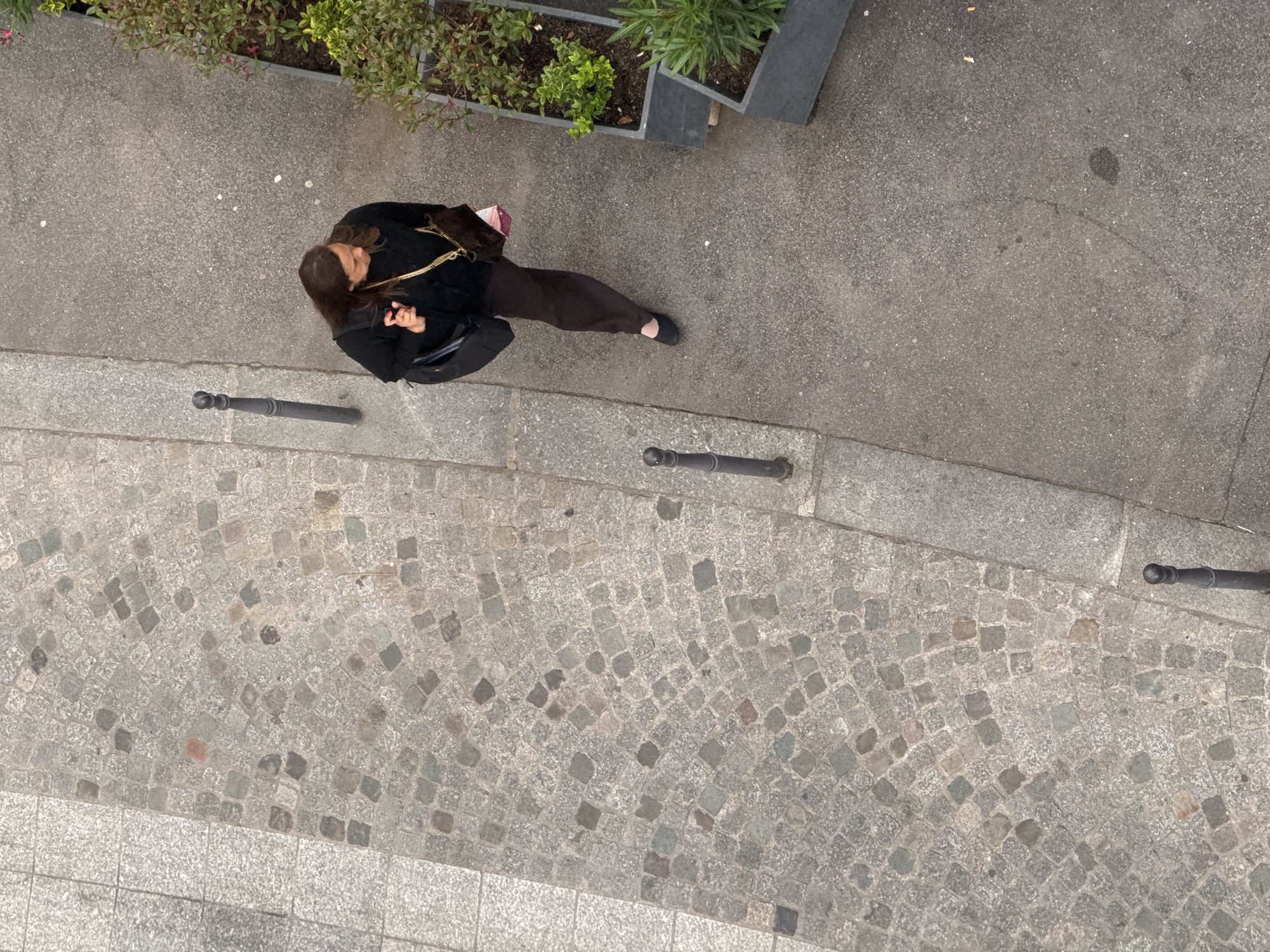 A high-angle shot shows a woman walking on a sidewalk in Paris, France. The woman has long brown hair and wears a black jacket and dark pants. She carries a black purse and a small pink bag. She appears to be looking at a cell phone in her hand. The sidewalk is adjacent to a cobblestone path, separated by short, black metal bollards. A planter with green plants is located along the top edge of the frame. The weather appears to be overcast.