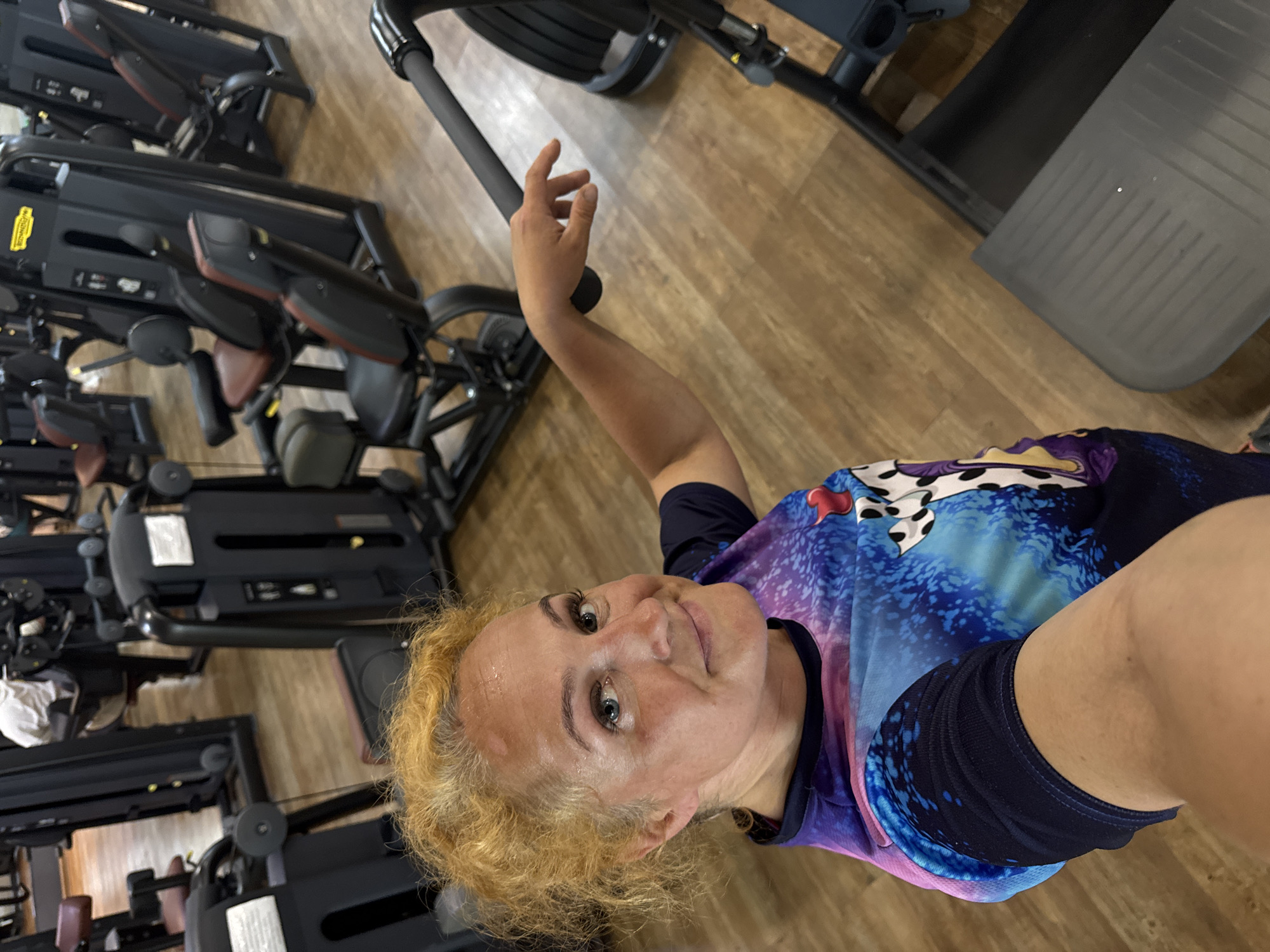 The image depicts a woman with blonde, curly hair and blue eyes taking a selfie in a gym. She is wearing a colorful t-shirt with a blue, purple, and pink abstract design and a black, long-sleeved shirt underneath. Her left arm is extended, and her hand is touching a black handle, likely from a gym machine. The background shows various gym equipment, including weight machines and what appears to be an elliptical trainer. The gym has wooden flooring. The woman is looking directly at the camera with a slight smile. There is no indication of the specific location within Chaikovsky, Russia.