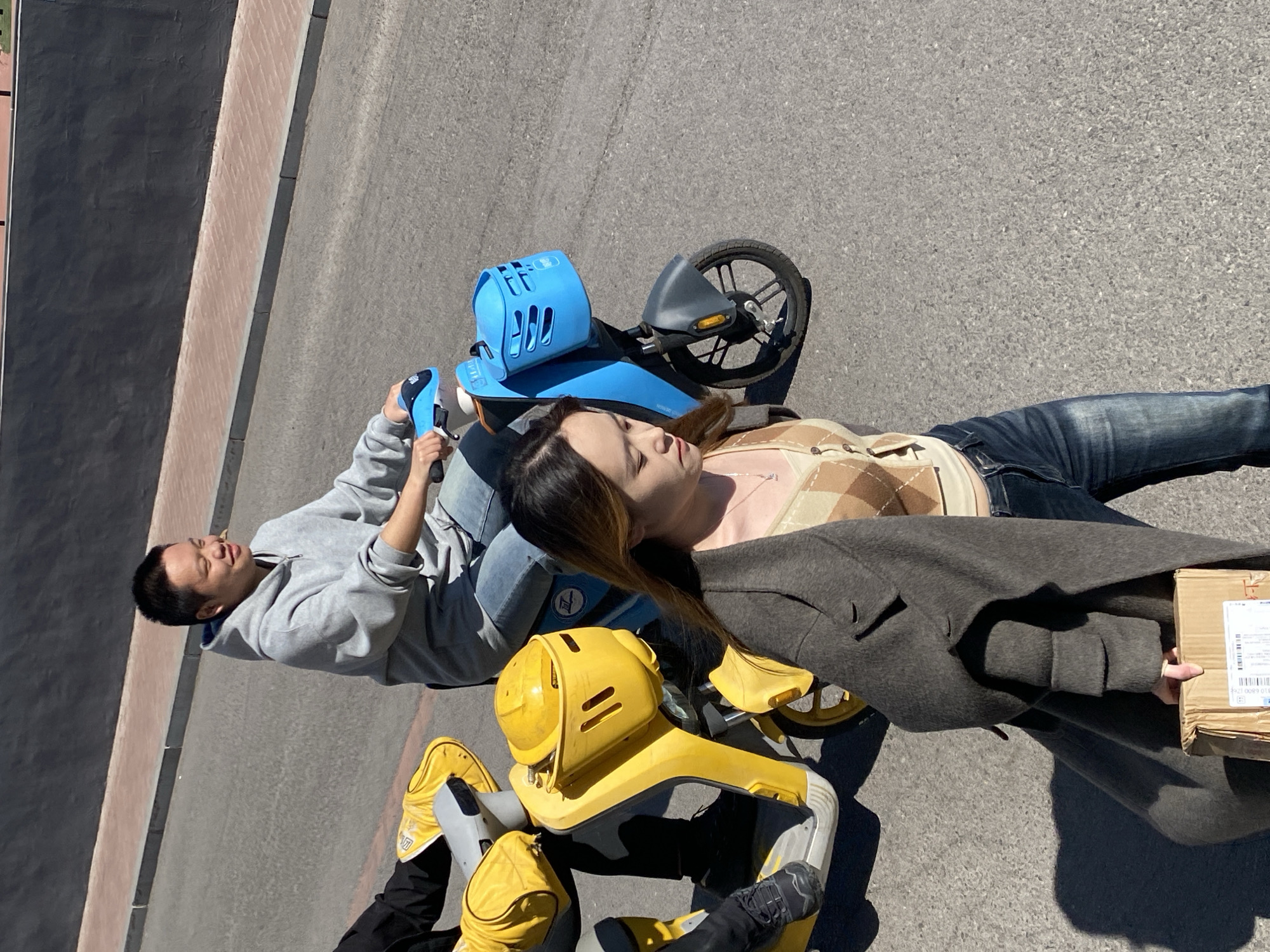 On an asphalt road in Zhongtun, China, under bright sunlight, three individuals are observed. In the foreground, an East Asian female with long, brown hair, wearing a light brown argyle-patterned crop top, dark blue jeans, and a dark grey coat, walks towards the right, carrying a brown cardboard box in her right hand. Behind her and slightly to the right, an East Asian male with short black hair, wearing a light grey hooded sweatshirt and blue jeans, smiles as he operates a light blue electric scooter equipped with a front basket. To the left and slightly behind the man, the lower body of a third person, wearing black pants and black shoes, is visible on a yellow electric scooter, which features a yellow front basket containing a yellow helmet. The road is bordered by a concrete curb and a dark grey wall in the background.