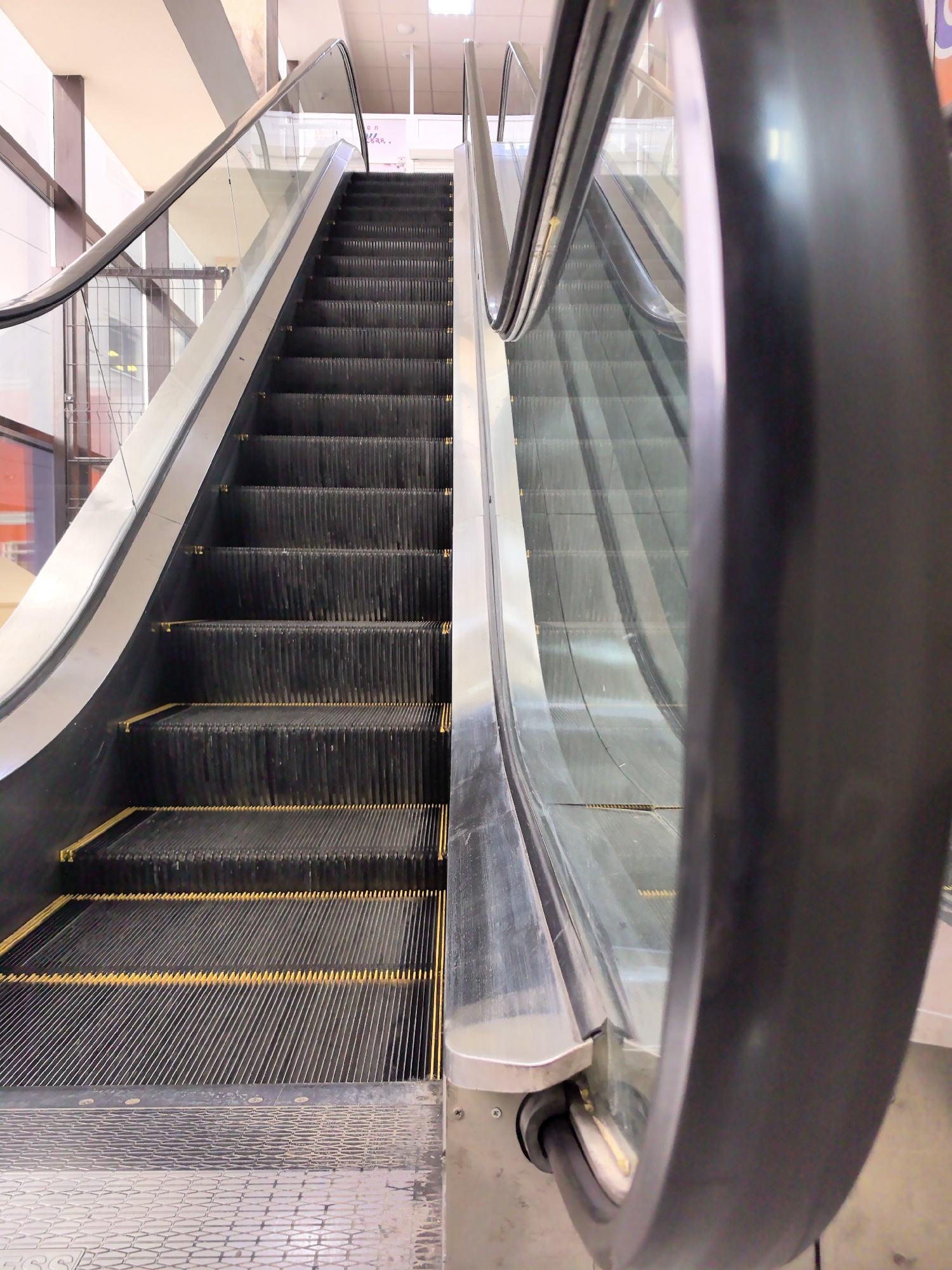 The image shows an escalator in an indoor environment. The escalator has black steps with yellow safety stripes and glass side panels with silver handrails. A black handrail moves alongside the steps. The escalator appears to be ascending, leading towards a lit ceiling. The surrounding architecture suggests the escalator is located in a commercial or public space, such as a shopping mall or transportation hub. The location is Unknown, Unknown.