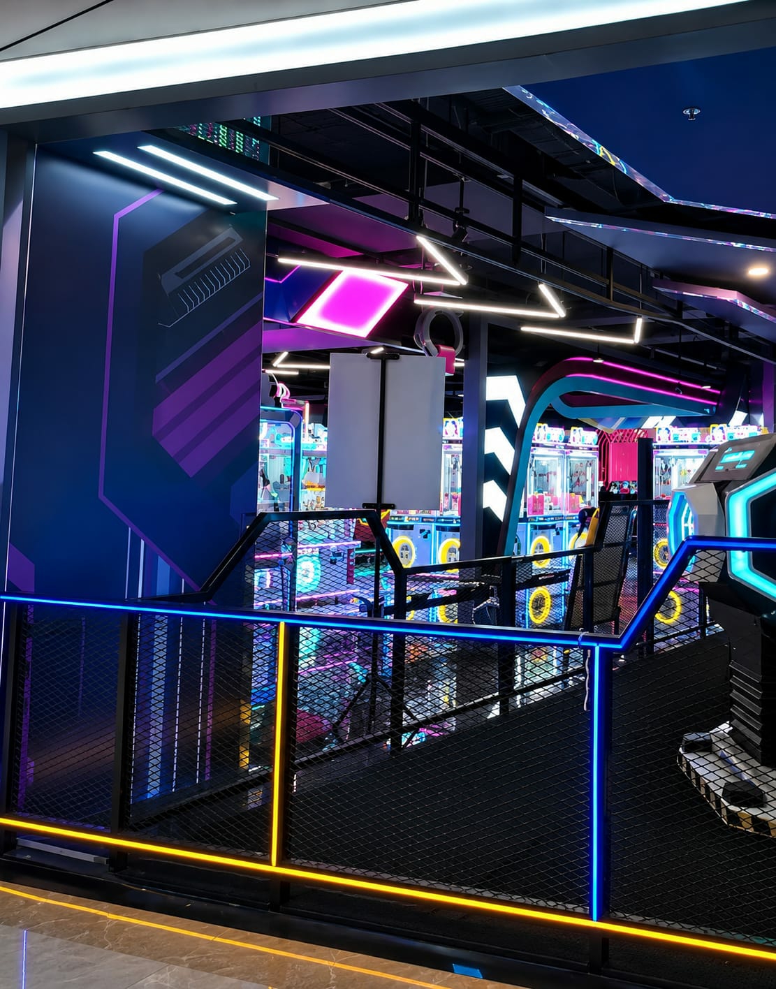 The image depicts the interior of an arcade or amusement center, likely located in the City of Medan, Indonesia, given the context provided. The environment is characterized by vibrant neon lighting, primarily in shades of blue, pink, and yellow, which illuminates rows of arcade games, claw machines, and other entertainment attractions. The foreground features a black metal mesh railing with blue and yellow neon lights outlining its edges, suggesting a walkway or elevated platform. Reflections of the colorful lights are visible on the polished floor. In the background, numerous glass-fronted game cabinets are arranged, many displaying colorful prizes or digital screens. The ceiling features a complex network of dark beams and pipes, interspersed with more geometric neon light fixtures. A large graphic with abstract purple and pink geometric shapes is visible on a dark wall to the left. The scene is devoid of people, creating a sense of anticipation and modern entertainment ambiance.