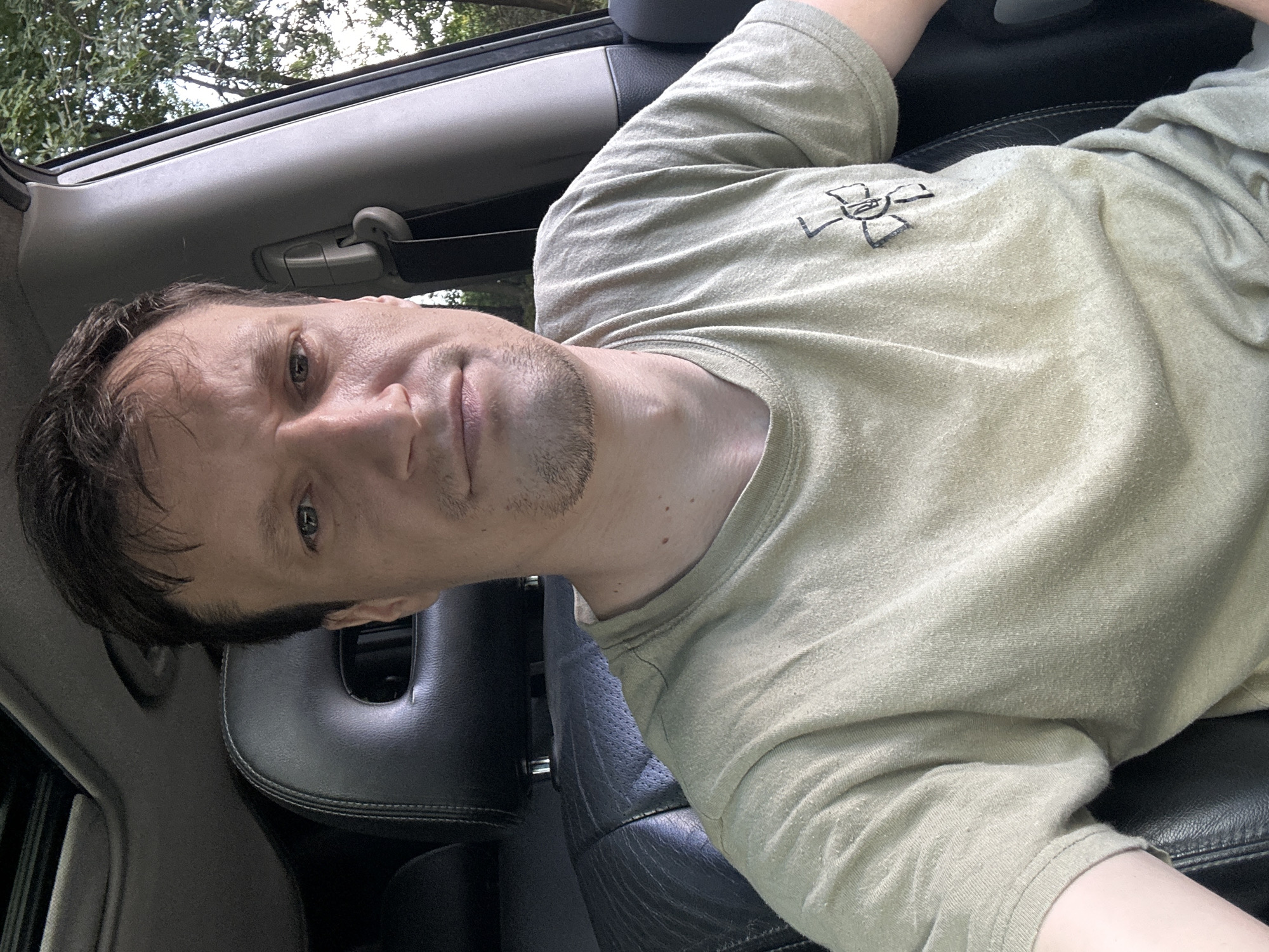 A man is sitting in the driver's seat of a vehicle, looking directly at the camera. He has short brown hair, a light beard, and is wearing a light green t-shirt with a symbol on the left chest. The interior of the car is visible, including a black leather headrest and seat. Through the open car door on the right, trees and foliage are visible, suggesting an outdoor setting. The image appears to have been taken during daylight. The metadata indicates the location as Vorobiivka, Ukraine.