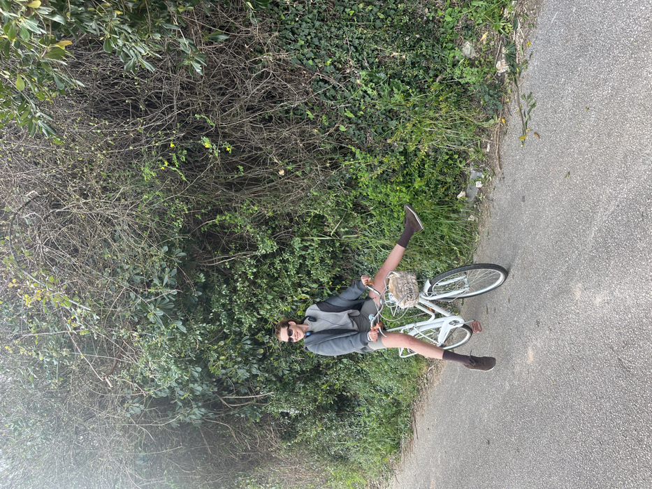 A single individual is observed on a white bicycle on a paved path in Budva, Montenegro. The subject, appearing female, wears sunglasses, a grey hooded jacket over a lighter grey top, olive-green shorts, brown socks, and brown shoes. Grey over-ear headphones rest around her neck. She is seated on the bicycle, holding the handlebars, with both feet lifted and extended horizontally outwards, suggesting a momentary levitation or pose. The bicycle is a white step-through model equipped with a front basket containing a light-colored woven object. To the right and rear of the subject is a dense wall of diverse green foliage, including shrubs, tangled dry branches, and groundcover, with occasional small yellow flowers. The paved path is grey asphalt. The scene is illuminated by natural daylight.