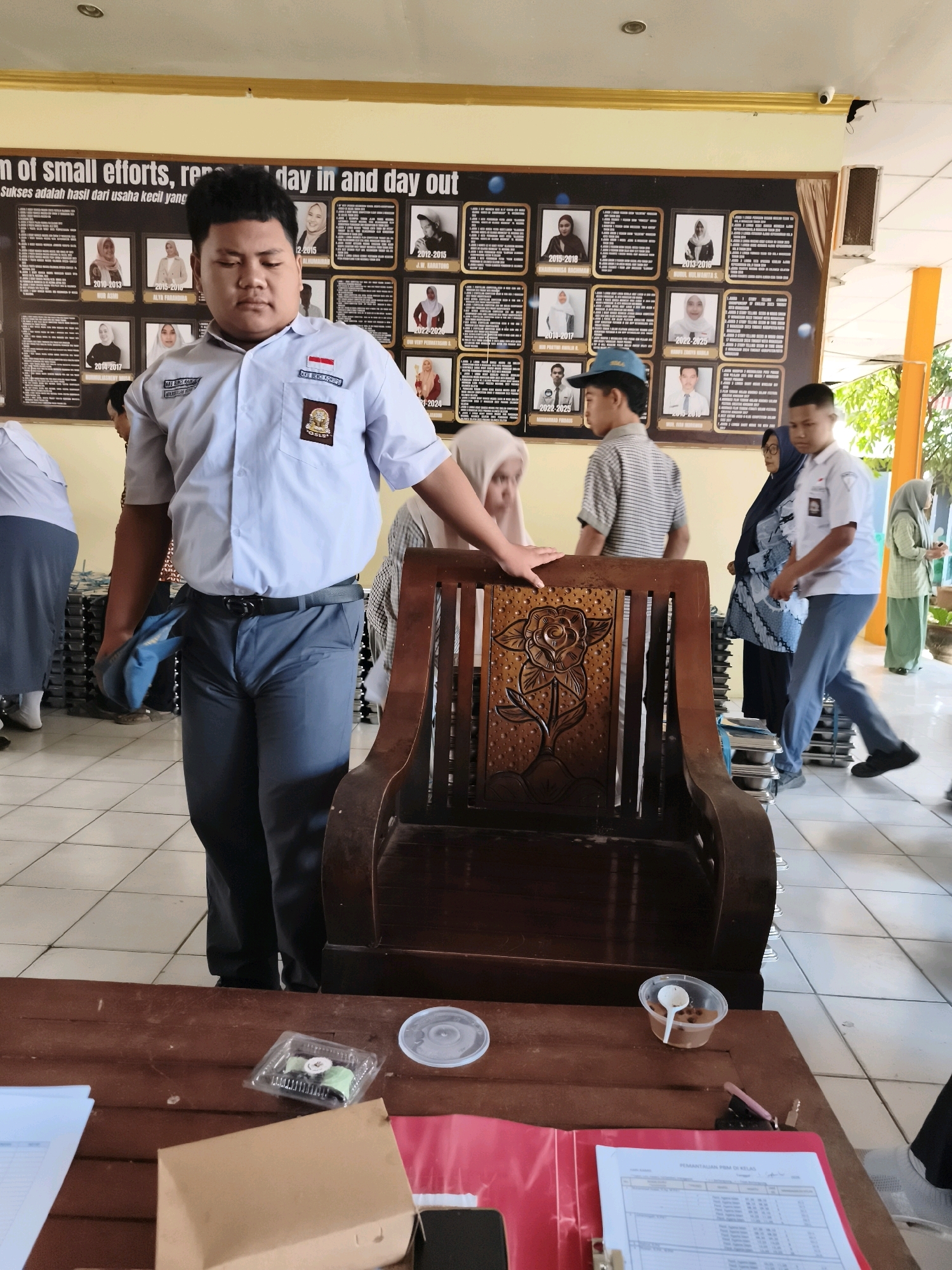 In an indoor setting, likely a school hallway or common area, a young person in a light blue uniform shirt and dark grey pants stands in the foreground, holding a folded blue cloth in their left hand. The uniform shirt features two patches on the left chest, one with a national flag and another with an emblem. Behind them, a large, ornate wooden chair with carved floral details is positioned centrally.
A wall display, a prominent feature in the background, showcases a grid of framed photographs, each accompanied by text. Some frames display portraits of individuals, while others contain text-heavy information. The display also includes the phrase "of small efforts, rep day in and day out" and "Sukses adalah hasil dari usaha kecil yang".
Several other individuals are visible in the background, also wearing school uniforms or modest clothing. Some are walking, others are standing, and one person is partially obscured by the wooden chair. The floor is tiled, and the walls are painted in light colors. A brown table is in the immediate foreground, with a clear plastic container holding a small pastry, a plastic cup with a spoon and some food, and a red folder containing documents. The presence of these elements suggests a school environment where students might be eating or engaged in academic activities. The style of the uniforms and the display on the wall suggest this is likely a school in Indonesia, and the mention of "Makassar" in the prompt, if accurate, places the scene within that specific Indonesian city.