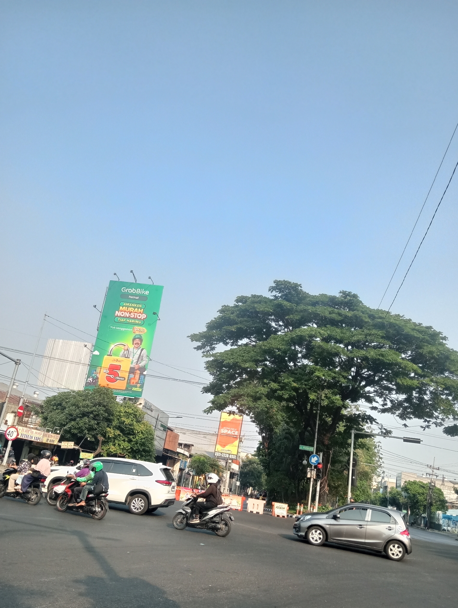 The image depicts a street scene in Surabaya, Indonesia, likely an intersection, characterized by a clear blue sky and bright sunlight. Several motorbikes and a white SUV are present on the road, with some motorbikes carrying passengers. A lone grey car is also seen driving on the right side of the road. In the background, a large green billboard for "GrabBike" is prominently displayed, featuring a graphic of a padlock and text in Indonesian. Adjacent to the billboard, a large, leafy tree dominates the upper right portion of the frame. Various buildings and traffic signals are visible along the street, suggesting a busy urban environment. The overall impression is of a typical day in a bustling Indonesian city.
