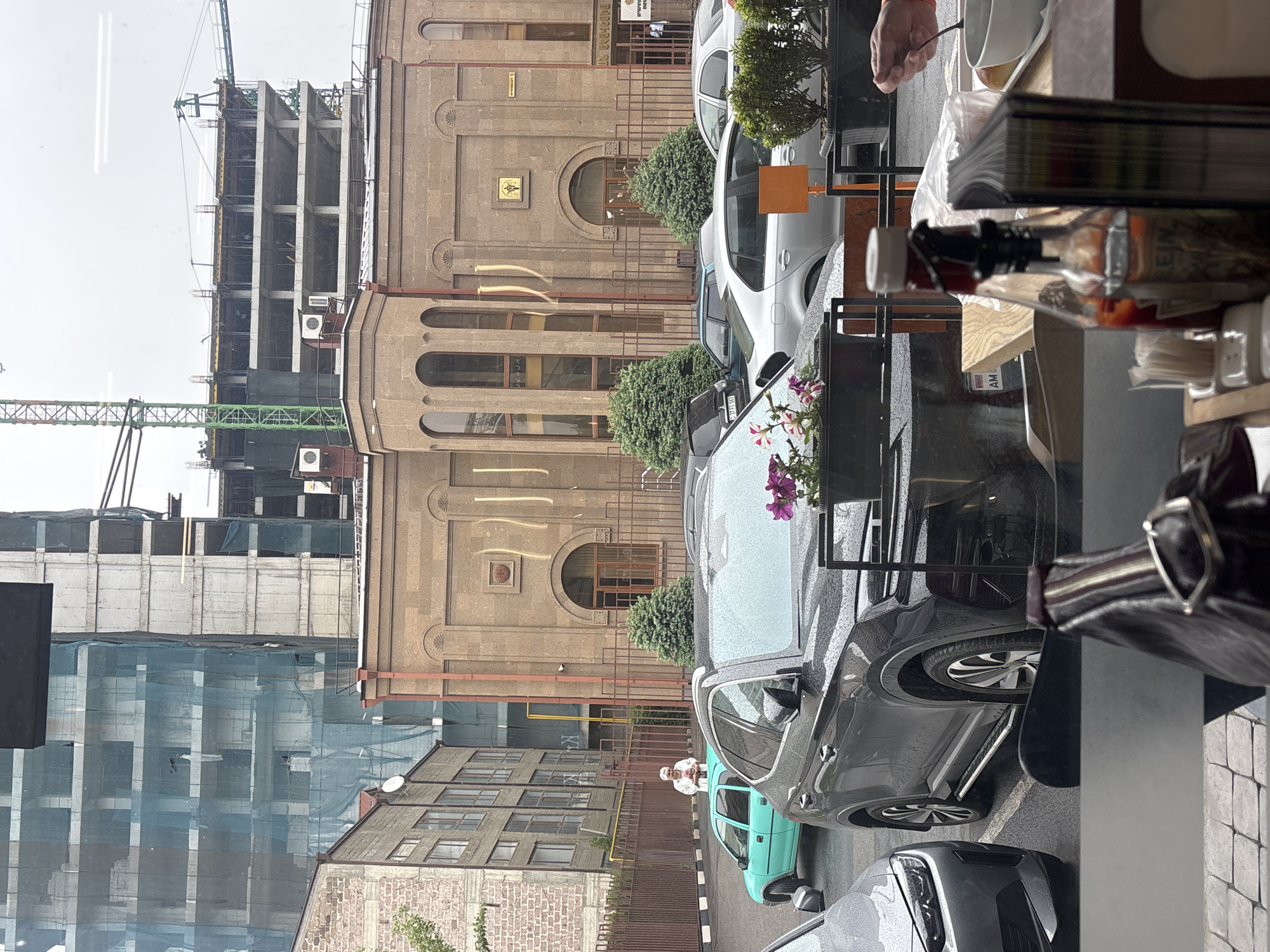 The image, taken from inside a building, captures an outdoor scene in Yerevan, Armenia. Through a glass window, there's a view of a street with parked cars. Prominent is a brown stone building with arched windows and decorative elements. To its left, a modern construction site features a tall building under construction, partially covered in protective netting. A crane is visible in the background. A smaller building with multiple windows sits in front of the larger buildings. Parked cars occupy the street. Foreground elements inside the building partially obscure the view.