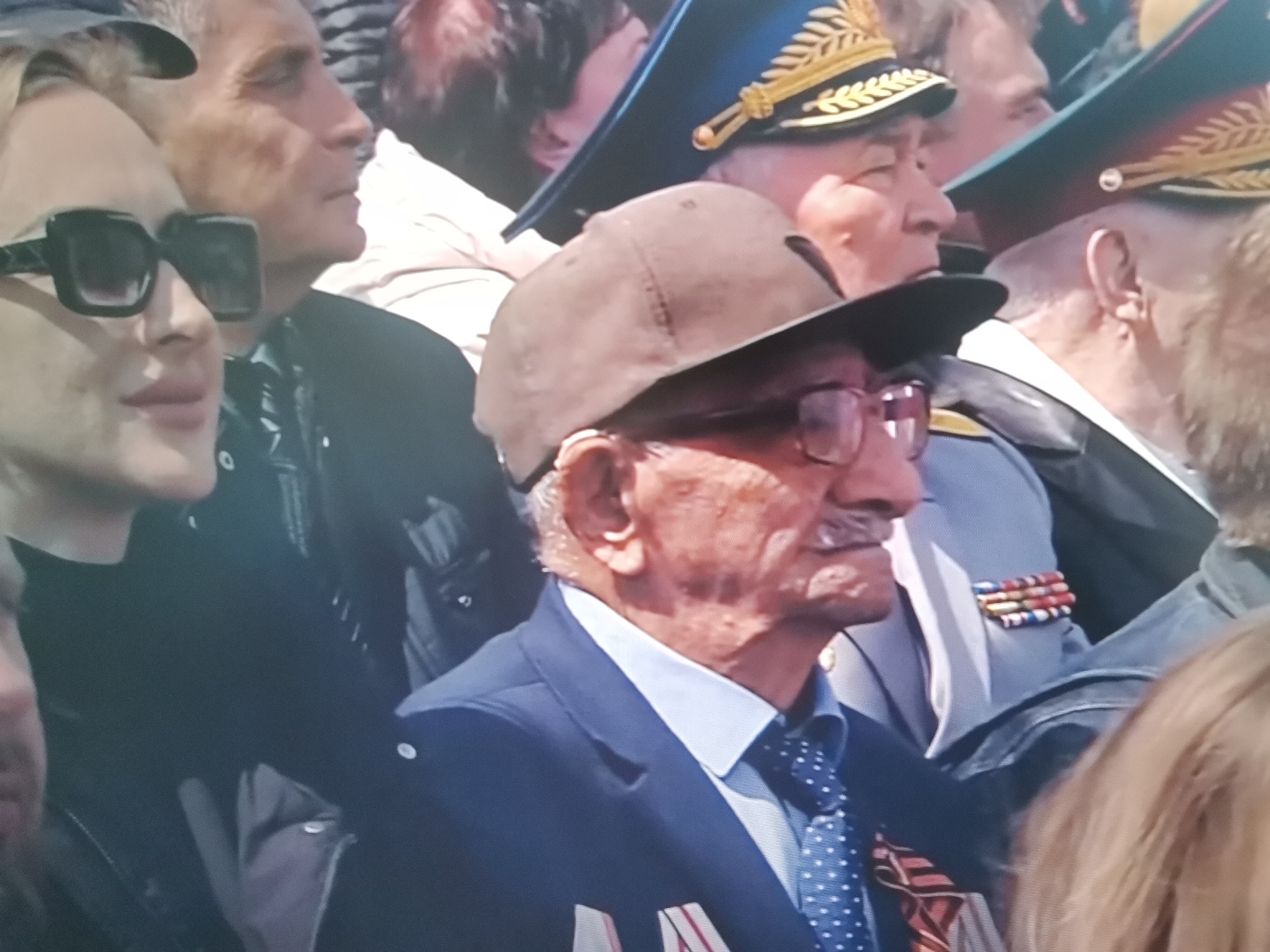 A medium shot, outdoor image captures a crowd of people in what appears to be a public event. In the foreground, a man wearing a dark blue suit, a light blue collared shirt, and a blue tie with white polka dots is visible. He has a light-colored baseball cap on and is wearing glasses. On his chest, he has a ribbon bar with multiple colored ribbons. To his left, a woman in sunglasses is partially visible. Behind the man in the suit, other individuals can be seen wearing military-style hats with gold trim. The overall impression is that of a formal gathering or parade. The location is identified as Belaazyorsk, Belarus.