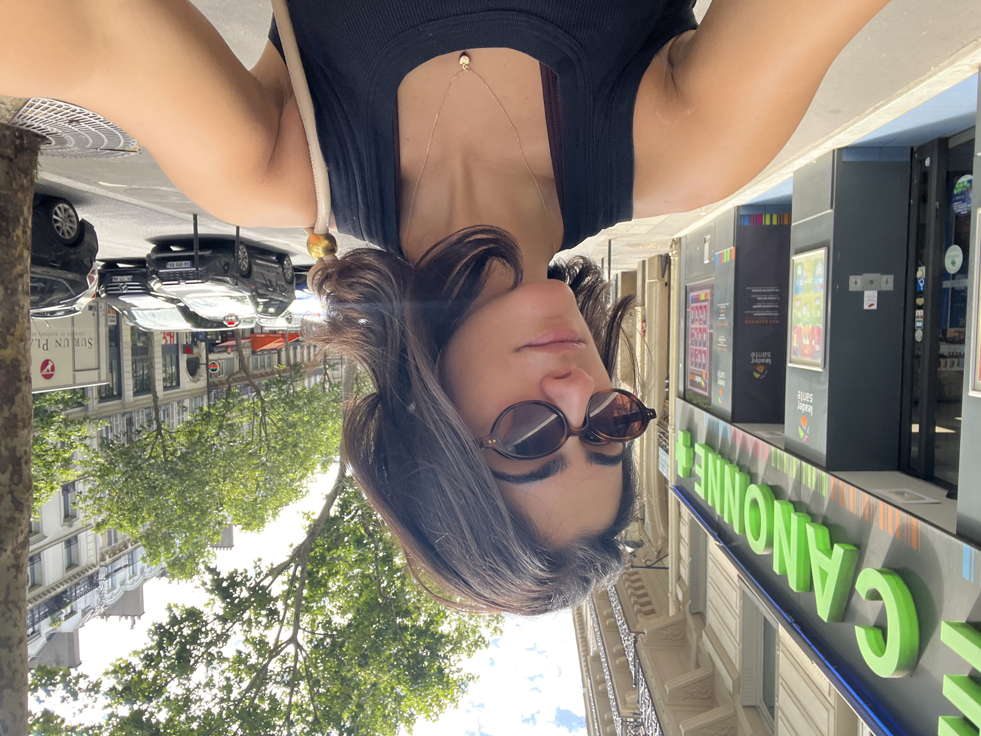 A woman with dark, shoulder-length hair and sunglasses takes a selfie while standing on a sidewalk in Paris, France. She is wearing a black tank top and a delicate gold necklace with a heart pendant. Behind her, a pharmacy sign with green lettering that reads "CANONNE+" is visible. The street is lined with trees and buildings characteristic of Parisian architecture. Several cars are parked or driving on the street, and a tree trunk is on the right side of the frame. The overall scene suggests a sunny day in an urban environment.