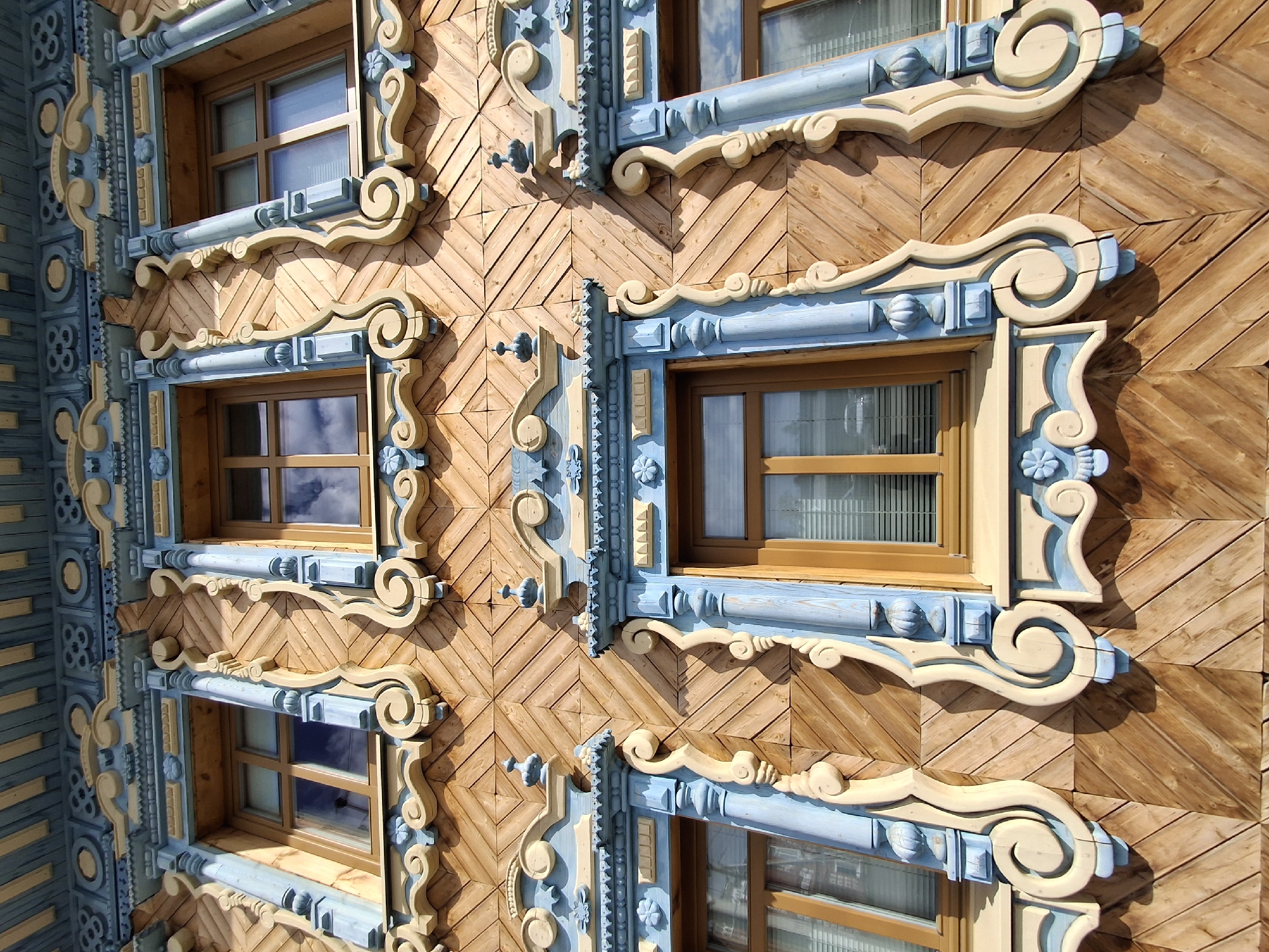 The image presents a detailed close-up of an ornate wooden building facade, characteristic of traditional Russian architecture, located in Sarapul, Russia. The facade is predominantly made of light-colored wood panels arranged in a diagonal, herringbone pattern, giving the wall a rich texture.
The most striking features are the intricately carved wooden decorative elements that frame the windows and create vertical architectural bands. These carvings are painted in soft shades of light blue and cream or beige, contrasting with the natural wood of the wall. The window frames are adorned with elaborate scrollwork, swirls, and stylized floral motifs. Between the windows, vertical sections display further decorative patterns, including geometric shapes like circles and squares, along with more scroll designs.
Multiple windows are visible, each framed by these decorative elements. The windows themselves appear to be wooden, with multiple panes, and some show reflections of the sky or clouds, suggesting clear weather. Light-colored curtains are visible behind some of the panes.
The scene is brightly lit by direct sunlight, casting strong, distinct shadows from the protruding carvings onto the wooden wall, enhancing their three-dimensional quality and suggesting it is daytime. There are no people or discernible activities taking place, and no visible text in the image. The focus is entirely on the craftsmanship and decorative artistry of the building's exterior.