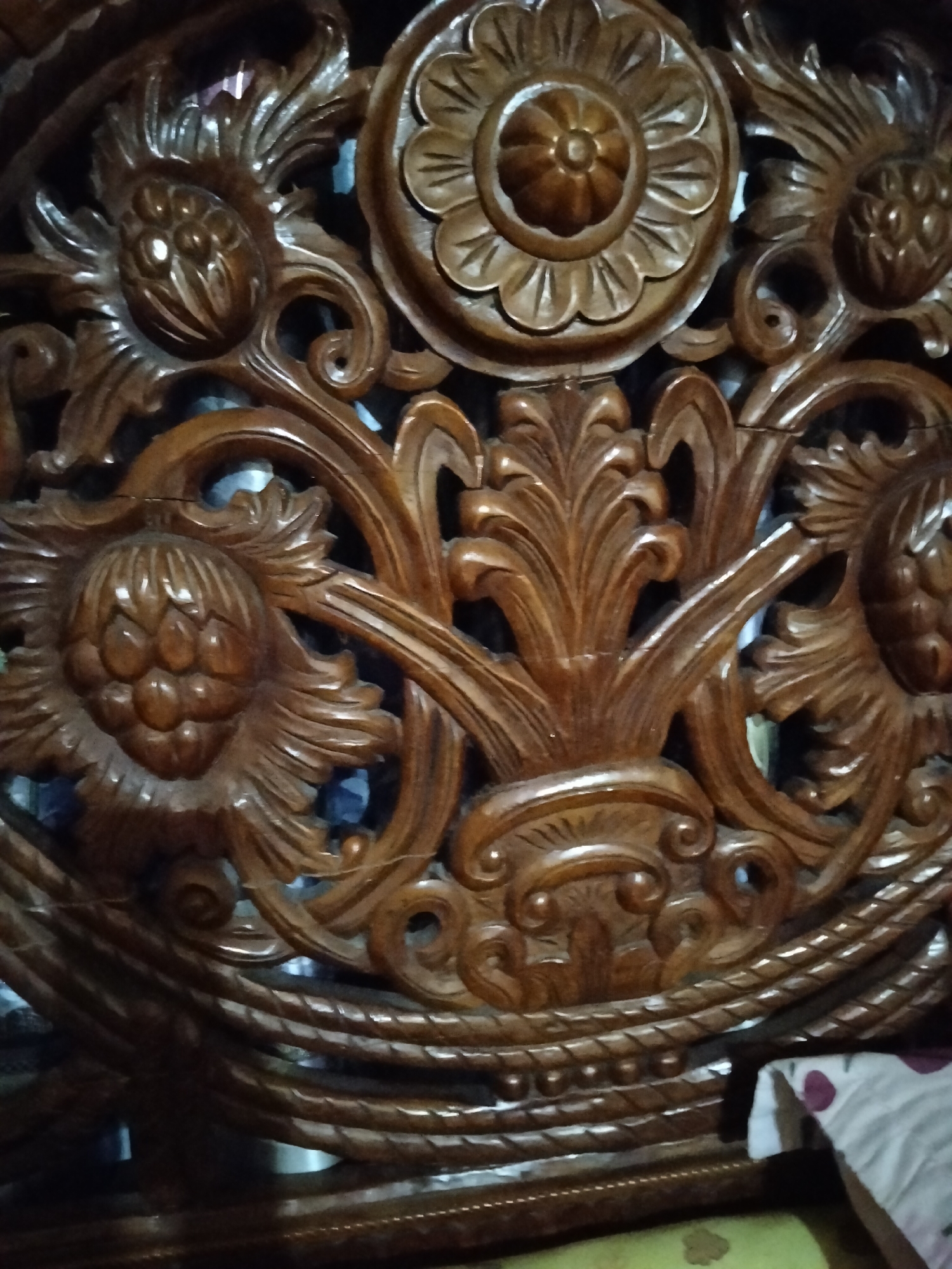The image shows a close-up of intricately carved wooden furniture, likely part of a bed frame or headboard. The carving features floral motifs, including a central circular design with petal-like extensions, and other elements resembling pinecones or clustered buds, all rendered with a high degree of detail and polished to a sheen. The wood appears to be a dark, rich brown, with visible grain and natural variations in color. The background is dark and out of focus, suggesting an indoor setting with low lighting. Part of a patterned textile, possibly a pillow or duvet, is visible in the lower right corner. The overall impression is one of craftsmanship and traditional artistry.
The location within Payagaboh, Indonesia, is not discernible from the image itself. This photograph captures a detail of an object and does not provide contextual environmental clues such as street views, landmarks, or specific types of buildings that would help pinpoint a precise location within the city.