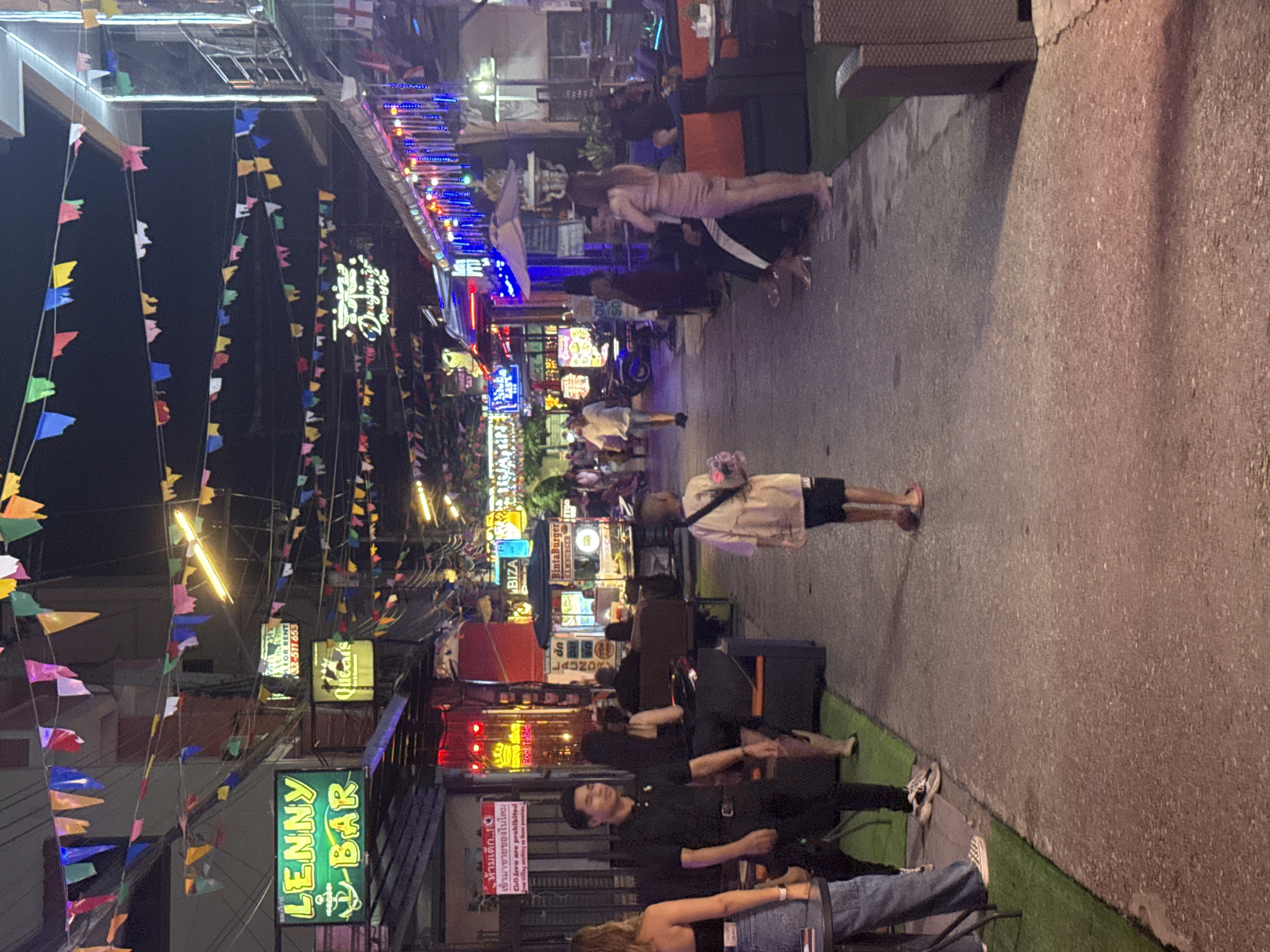 The image depicts a bustling street scene at night in Hua Hin City Municipality, Thailand, likely a tourist-oriented area with numerous bars and food stalls. Colorful decorative flags are strung across the street, creating a festive atmosphere. The street is paved, with artificial turf bordering one side.
Several individuals are visible:
- On the left, a woman in jeans and a black top is partially visible.
- A man in a black polo shirt and dark pants is walking away from the camera.
- A young boy in a light-colored shirt and dark shorts is walking towards the camera.
- To the right, a woman in a pink dress is standing and conversing with someone seated. Several other individuals are seated at tables or in lounge chairs, appearing to be patrons of the establishments.
Various businesses are identifiable by their signage:
- "LENNY BAR" is prominently displayed with a neon green sign.
- A sign for "Queer's" is visible.
- A sign for "BIZA" and "Binta Burger" indicate food vendors.
- Other signs for bars and services are present but less legible.
The environment suggests an active nightlife scene, with illuminated signs and people engaging in social activities. The overall impression is one of a lively commercial street in a Thai resort town at night.