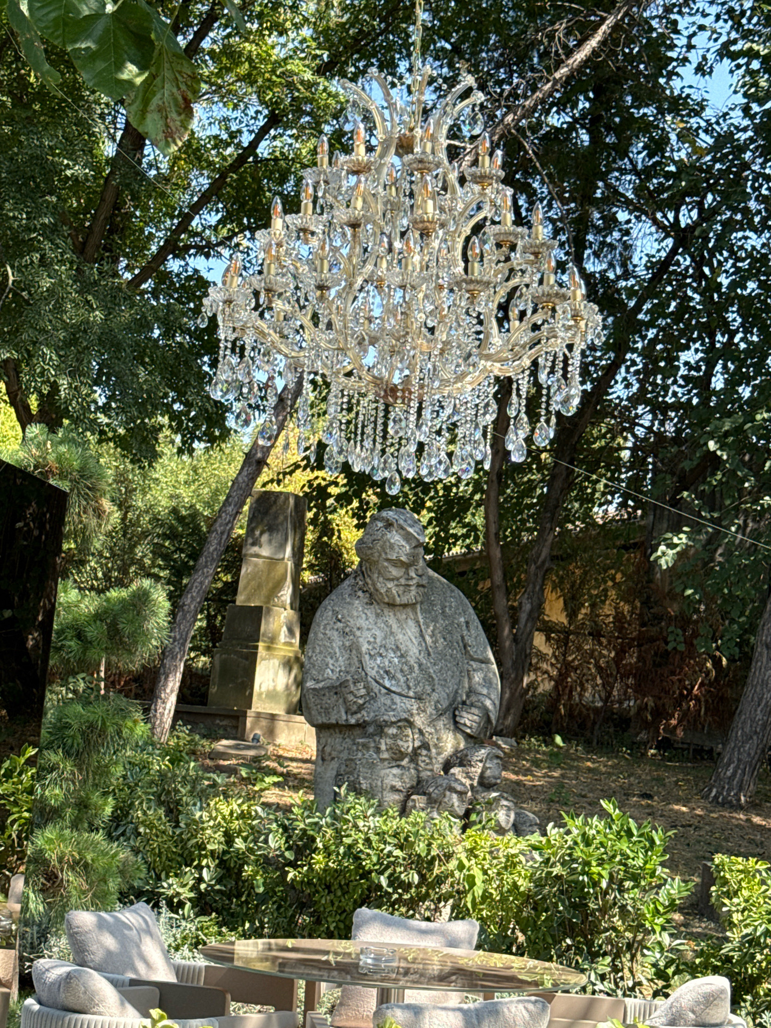 A large, ornate crystal chandelier hangs from a tree branch, suspended above a stone sculpture of a seated figure in a garden setting. The sculpture appears to be of a person with a beard, wearing a cap and coat, with smaller figures at their feet. Behind the sculpture, a tiered stone structure is visible, possibly a monument or part of a cemetery. The garden is lush with greenery, including trees, bushes, and pine trees, indicating an outdoor location. In the foreground, outdoor furniture, including chairs with cushions and a glass-topped table, suggests a seating area. The overall scene is one of juxtaposition, with the formal elegance of the chandelier and sculpture set against the natural backdrop of a garden. The lighting suggests it is daytime, with sunlight filtering through the trees. The specific location within Bucharest, Romania is not discernible from the image itself.