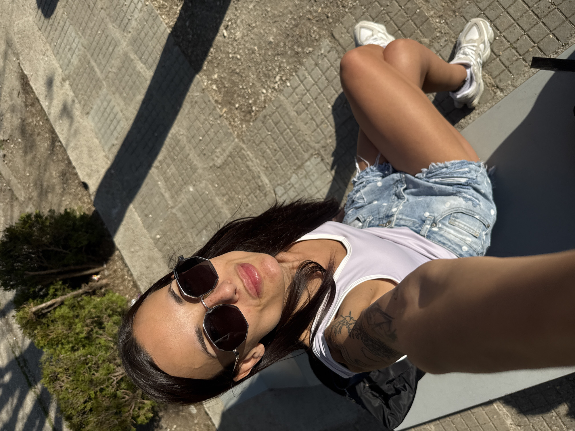 A woman is taking a selfie outdoors. She is wearing sunglasses, a white tank top, and denim shorts. She is sitting with her legs crossed and wearing white sneakers. Her left arm is visible and features a tattoo. She is sitting on a gray ledge, with paving stones and concrete visible in the background. There is a small tree behind her. The location is likely in Pleven, Bulgaria, based on the user's query.