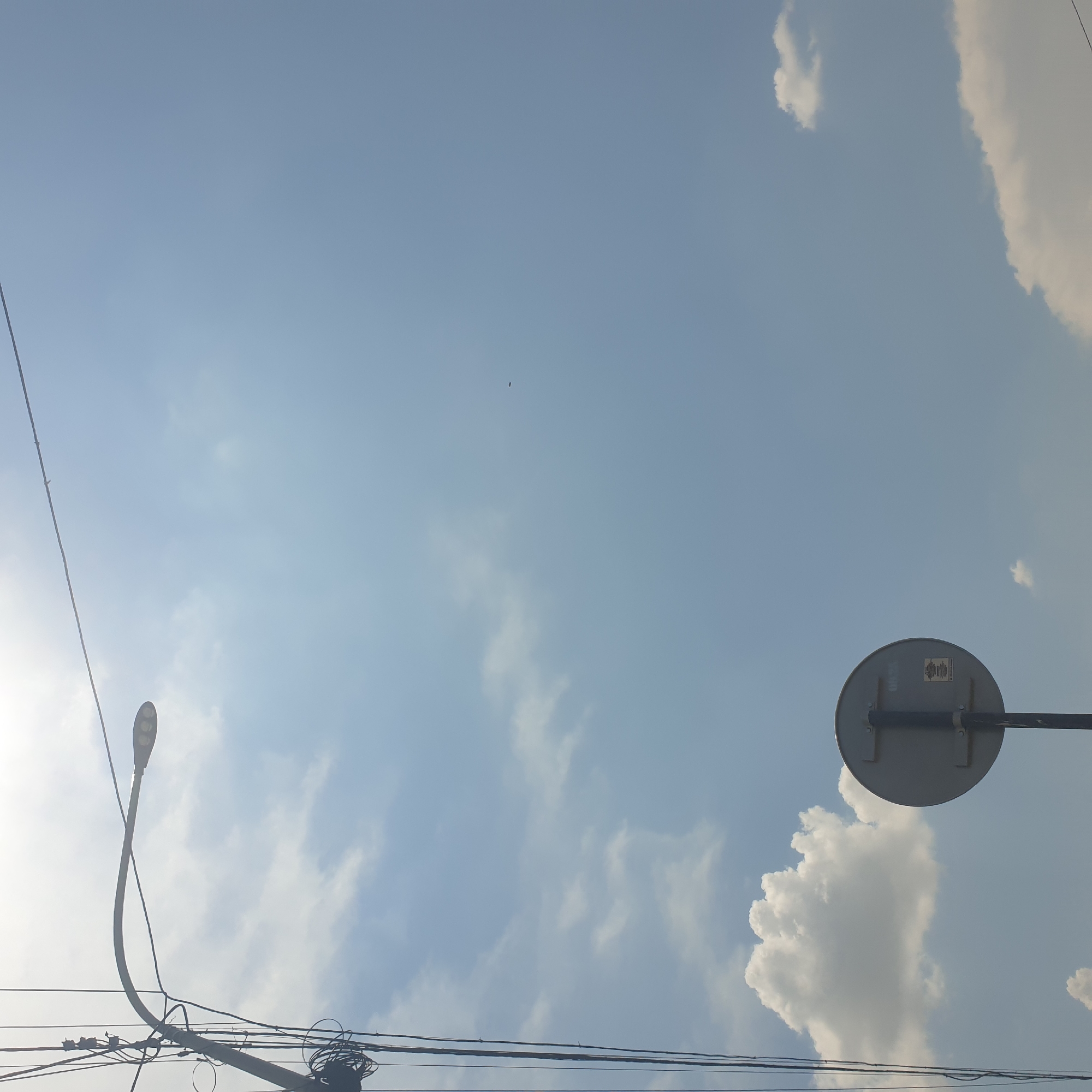 The image is a low-angle outdoor shot, likely taken in Shymkent, Kazakhstan, focusing on the sky with a lamppost and a traffic sign. The sky is a pale blue with scattered white clouds. A modern-looking lamppost with multiple LED lights arcs from the left side of the frame. Numerous electrical wires are tangled around the lamppost's pole, creating a complex network. In the lower right quadrant, a round, grey traffic sign is mounted on a dark pole, facing away from the camera. The back of the sign has some text and a sticker. A small, dark, indistinct object, possibly a bird, is visible in the upper right portion of the sky. The overall scene depicts an urban sky with typical infrastructure elements.