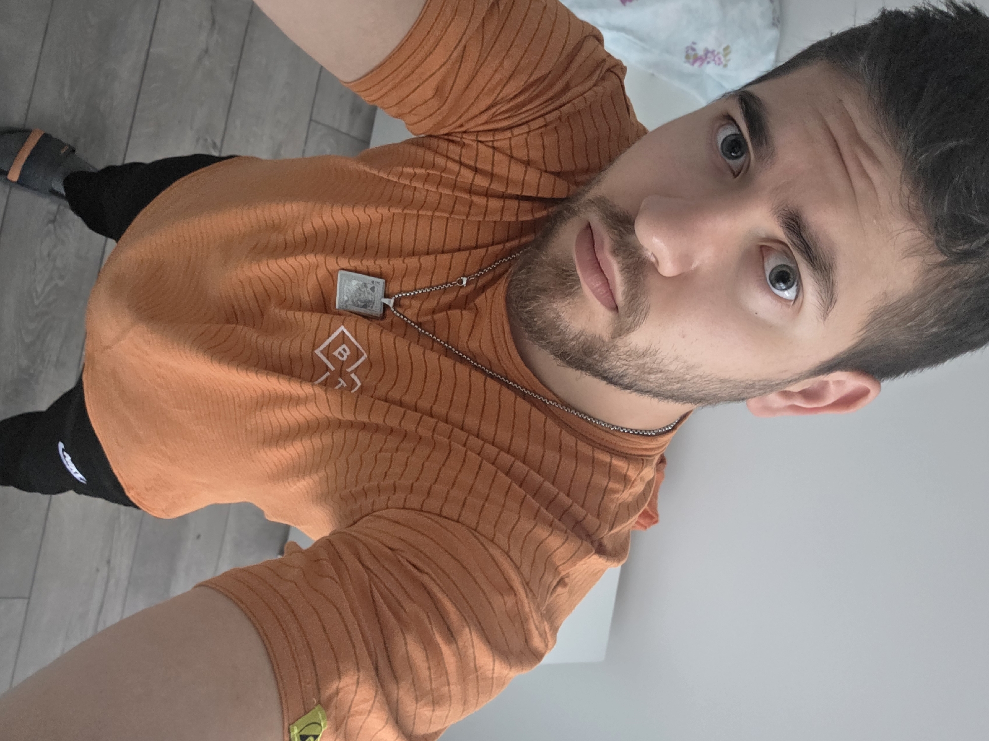 A man wearing an orange t-shirt, black pants, and grey sandals is taking a selfie. He is indoors, standing on grey flooring. A necklace with a square pendant is visible. The location cannot be determined to be Yagodovo, Bulgaria from the image.