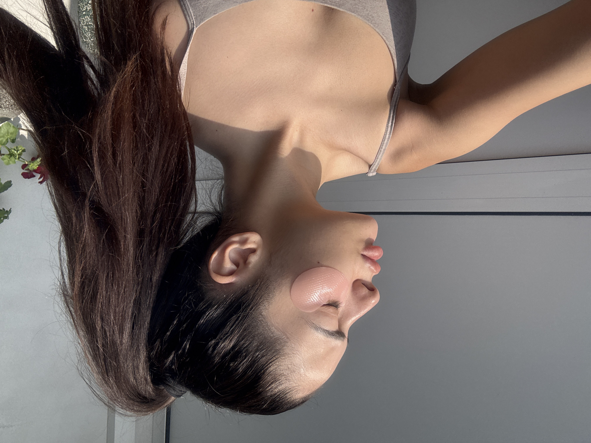 A profile portrait captures a young individual, likely female, engaged in a self-care routine indoors. The image is rotated 90 degrees counter-clockwise, with the person's head positioned on the right, looking slightly upwards. They have long, dark brown hair styled away from their face, flowing towards the left. Two translucent, pink hydrogel under-eye patches with a subtle textured surface are visible beneath their closed eyes, indicating a moment of relaxation or skincare. Their lips are slightly parted.
The individual wears a light grey tank top or camisole, revealing their upper torso and arm. Strong, directional sunlight illuminates the scene, casting prominent shadows across their neck, collarbone, and arm, suggesting it is either morning or late afternoon. The warm light highlights the smooth texture of their skin.
A small plant with green leaves and a hint of red blossoms is partially visible on the far left of the frame. The background consists of a plain, light grey wall or interior surface, with a white window frame visible near the bottom edge (if the image were upright). The overall atmosphere is serene and calm, capturing a quiet moment of self-care taking place in Plovdiv, Bulgaria. No text is visible in the image.