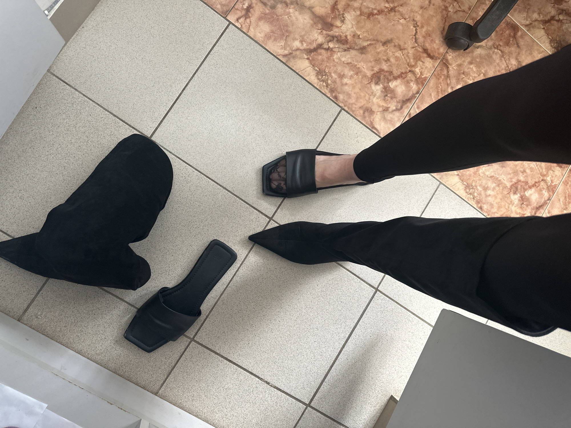 This high-angle shot provides a detailed view of an indoor tiled floor in Chaikovsky, Russia, focusing on a person's lower legs and several footwear items. The floor is predominantly covered with light gray, speckled square tiles featuring dark grout lines, which meet a diagonal section of brownish-pink marble-patterned tiles in the upper right.
A person's right foot, clad in a black slide sandal with a square toe and a sheer black patterned (possibly lace) stocking, extends from the right side of the frame. This foot is accompanied by a leg dressed in black pants. To the left, on the light gray tiles, another identical black slide sandal rests beside a black, soft-material shoe, possibly a boot or slipper with a pointed toe, lying on its side.
Bright lighting, likely from a window, casts distinct shadows across the floor and the person's leg, indicating it is daytime. In the upper right, a black caster wheel from an office chair is partially visible on the marble-patterned tiles. A small corner of a light gray, flat surface, potentially a desk or table, is seen in the bottom right. The scene suggests a moment of changing shoes or having them off while indoors, likely in a home or office setting. There is no visible text in the image.
