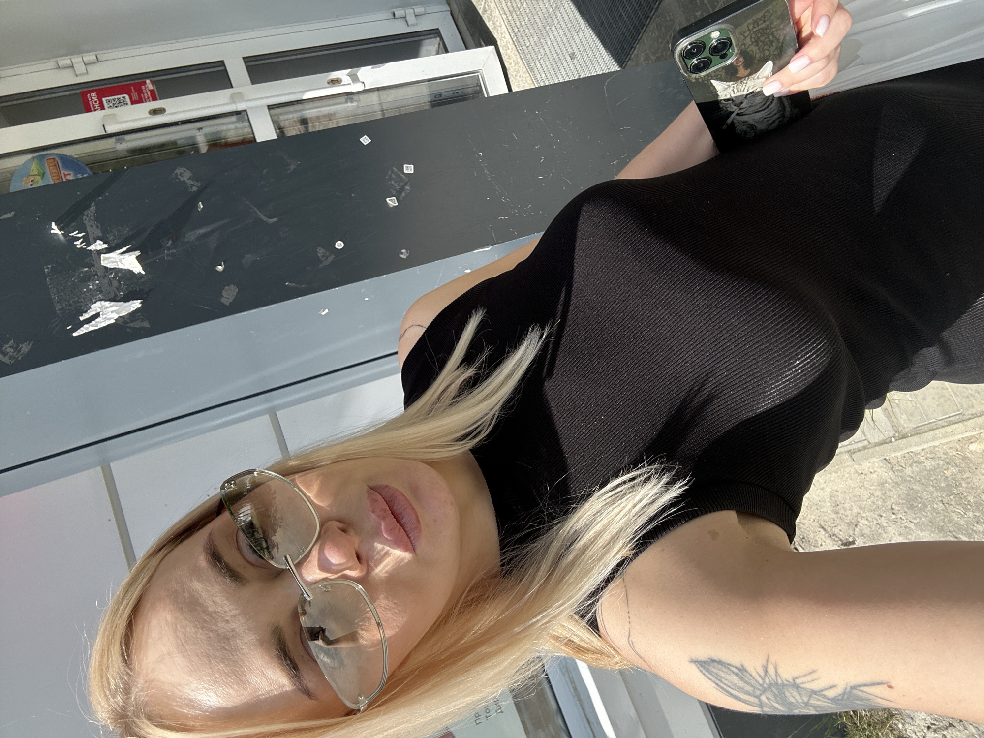 An adult female with long blonde hair and fair skin is captured in a sunlit selfie, standing outdoors next to a building facade in the city of Unknown, Unknown. She wears large, reflective, rimless or semi-rimless sunglasses with light-colored lenses, partially obscuring her eyes. Her lips appear full. She is dressed in a form-fitting, ribbed, sleeveless black top or dress. A prominent black-ink tattoo, depicting an insect (possibly a cicada or large fly), is visible on her left upper arm. In her right hand, she holds a modern smartphone featuring a clear case with an image of a feline and a triple-lens camera module; a small white bag or object is also held in the same hand.
The immediate background consists of a dark grey/black vertical panel of the building, showing various scuff marks and remnants of small white stickers. To the right, a white-framed glass door is visible, through which a red sign with a QR code and the Cyrillic word "АКЦИЯ" (meaning "SALE" or "PROMOTION") can be seen. Another partially visible round sign with a cartoon baby and the Cyrillic word "ОТ" is also visible through the glass. A dark grey corrugated doormat is situated on the pavement outside the door. The ground is concrete pavement with sparse dry vegetation. Strong shadows indicate direct sunlight.
