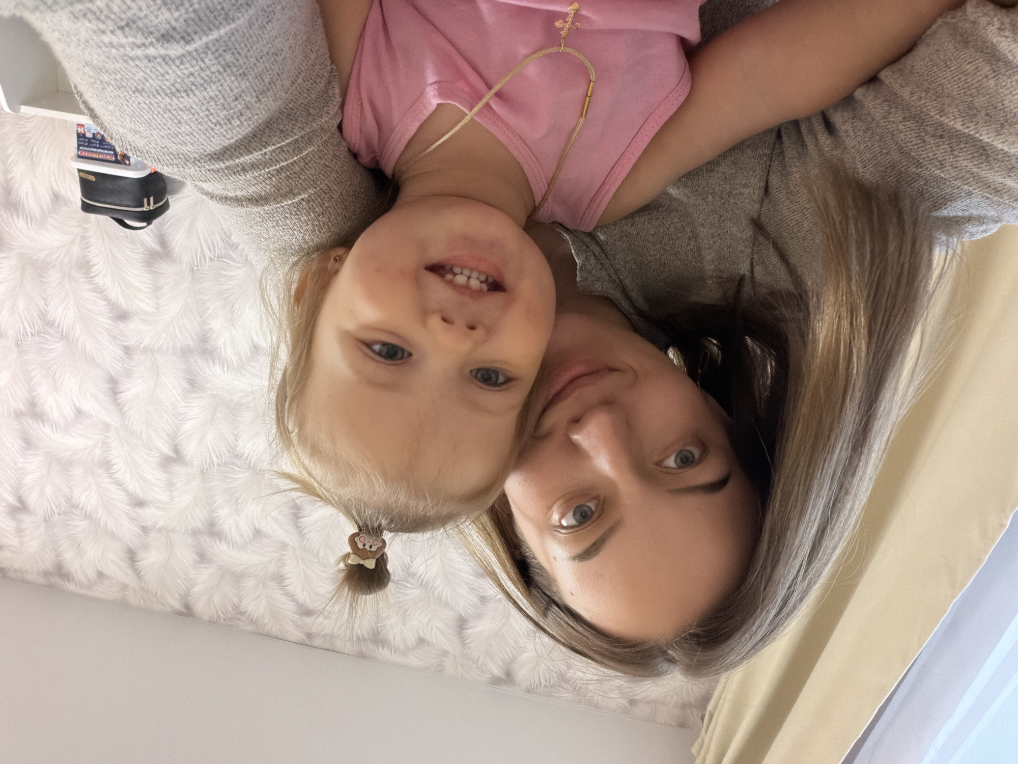 A woman and a young girl are close together, facing the camera. The girl wears a pink shirt and a necklace with a cross. The woman has long, light brown hair and is wearing a gray top. The background includes a textured, white wall and a curtain. An object is visible on a white shelf in the upper left corner. The image is taken indoors. The location is Reshetnikovo, Russia.