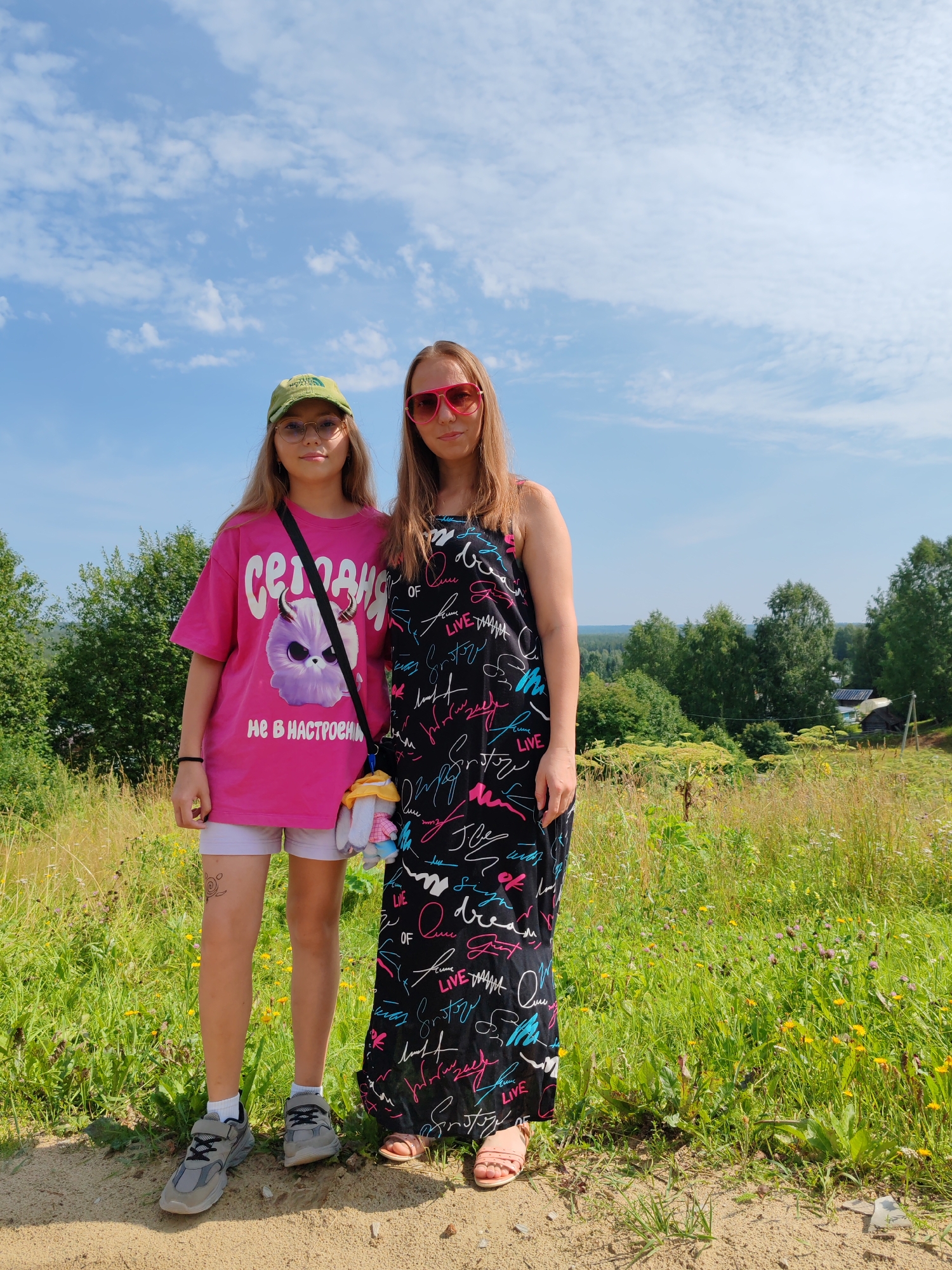 Two individuals, likely a younger and older female, are standing on a dirt path in a grassy, rural area with trees and a distant view of buildings under a partly cloudy sky. The younger female on the left is wearing a pink t-shirt with a graphic of a cartoon creature and text, white shorts, a green baseball cap, glasses, and grey sneakers. She has a black crossbody bag with a plush toy attached. The older female on the right is wearing a long, black dress with a colorful, handwritten-style pattern and pink sunglasses. She is wearing pink sandals. The environment appears to be a scenic overlook with lush green vegetation and a slightly hazy horizon. The presence of dirt paths, grassy fields, trees, and simple buildings suggests a non-urban, possibly village or countryside setting. No specific landmarks or signs indicate the city of Чухлэм, Russia.