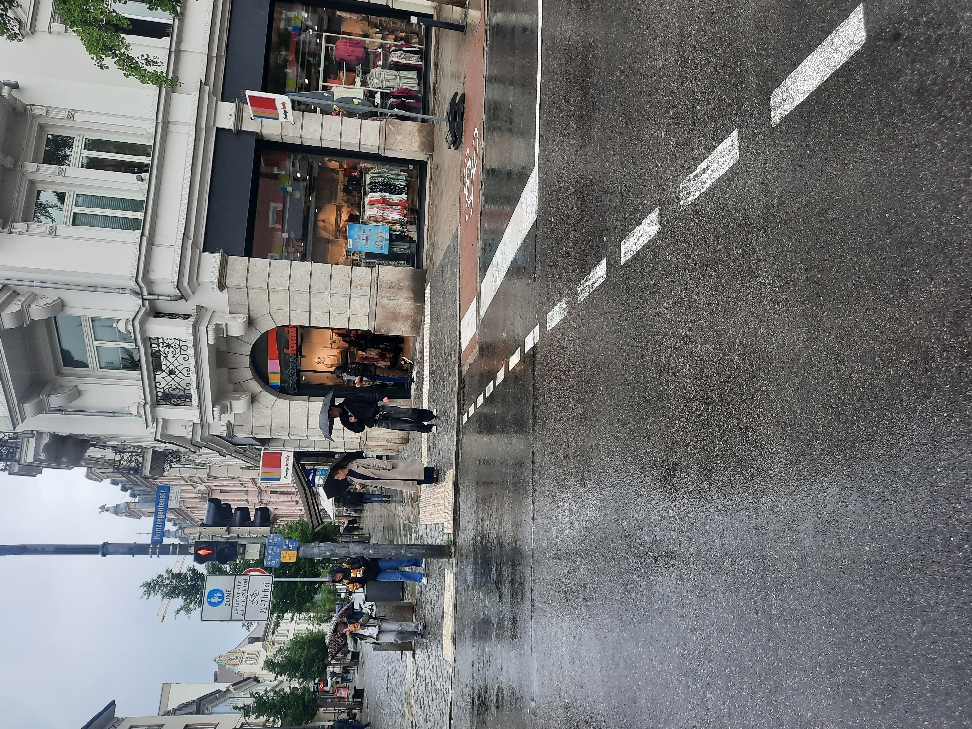 The image depicts a street scene in Rosenheim, Germany, on a rainy day. The road is wet and reflects the surrounding buildings and streetlights. Several people are present, most of whom are holding umbrellas.
On the left side of the street, a building with several windows and ornate balconies is visible. In front of it, a traffic light is red, indicating that vehicles should stop. Adjacent to the traffic light are several street signs, including a "ZONE" sign with pedestrian and bicycle symbols, and a sign indicating delivery traffic restrictions.
Several individuals are standing on the sidewalk, some under umbrellas, waiting to cross the street. One person is holding a shopping bag. Further down the street, a store with large display windows is visible, with mannequins showcasing clothing. The store appears to be a clothing retailer.
The overall atmosphere is one of a typical European city street during inclement weather. The wet asphalt, umbrellas, and muted lighting contribute to the scene's somber mood. The presence of the store and the street signs help to establish the urban setting.