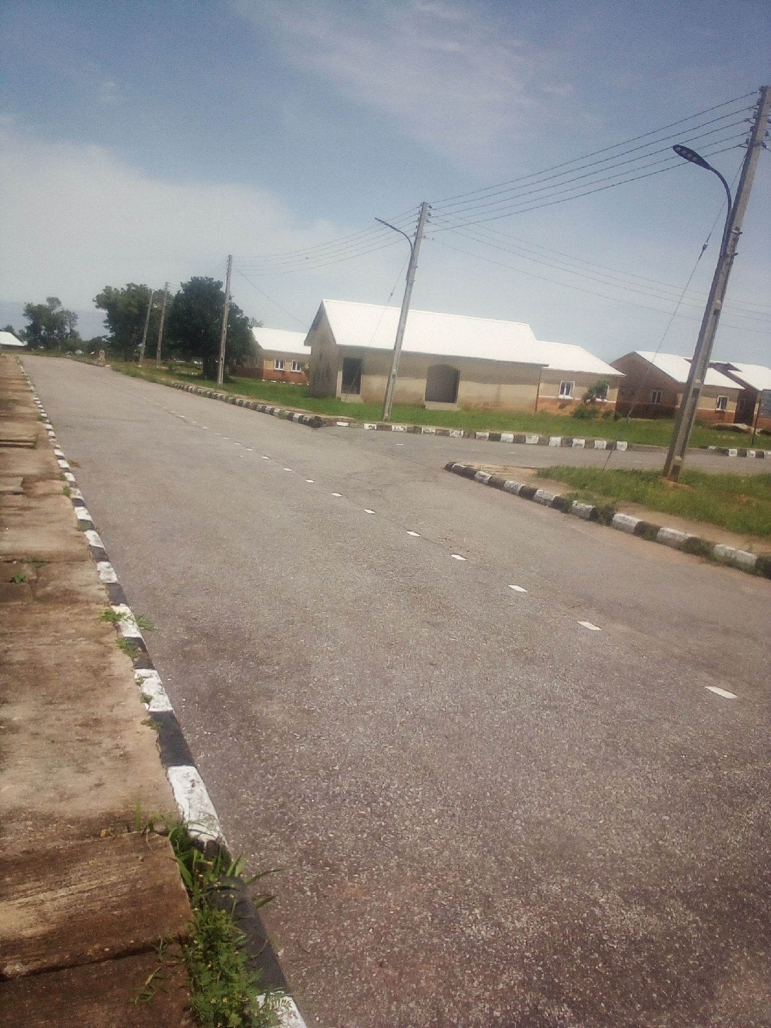 The image depicts an asphalt road in the city of Jauro Yinu, Nigeria, lined with concrete curbs that are painted with black and white stripes. The road is bordered by a strip of grass and vegetation. On the right side of the road, there are several single-story buildings with white roofs, appearing to be residential or administrative structures. A utility pole with power lines is visible next to the road, and another pole is seen in the distance. The sky is partly cloudy and blue. The scene appears to be a quiet, possibly newly developed or maintained area within the city, with no people or vehicles visible.
