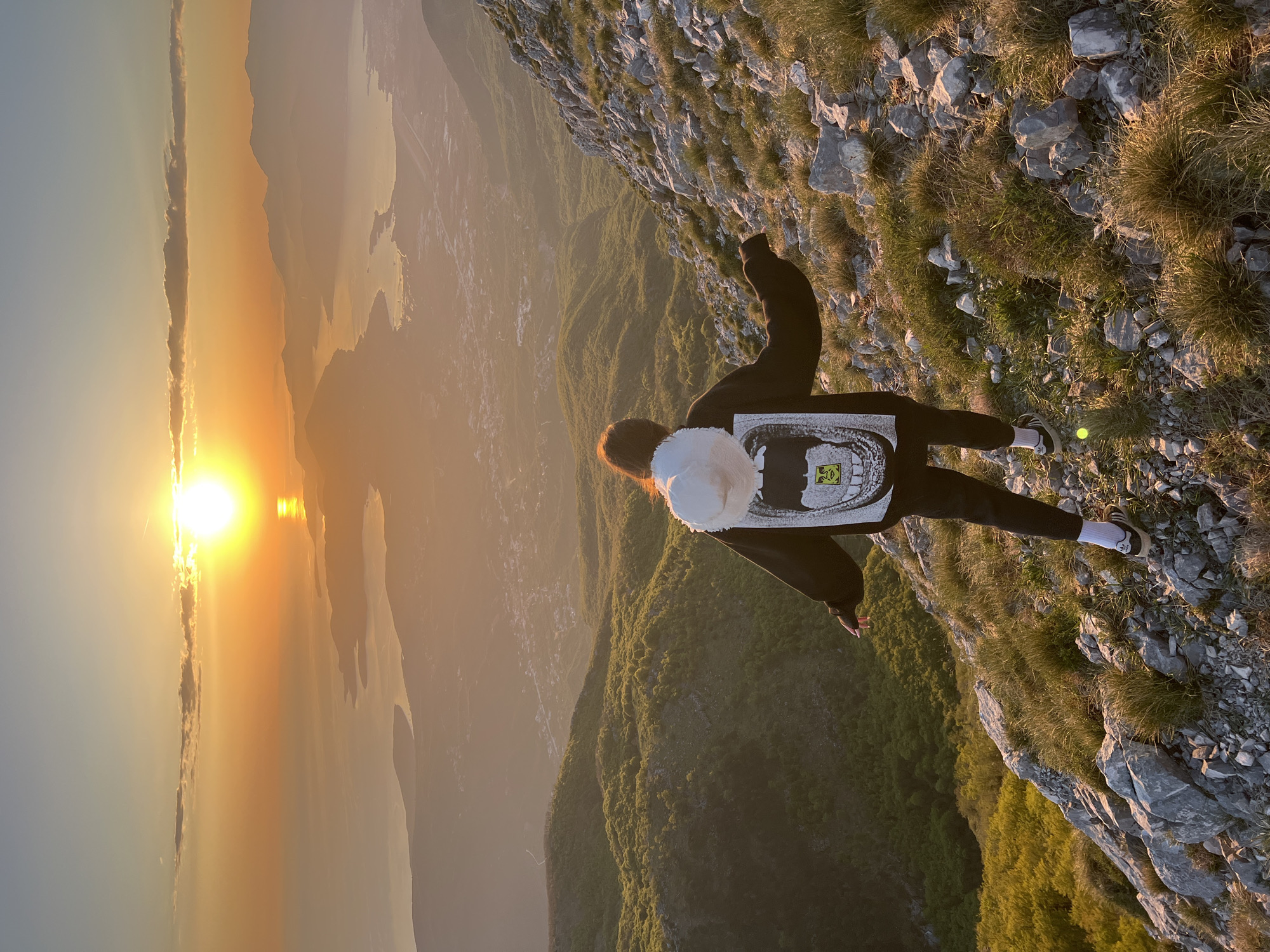 A person is standing on a rocky cliff edge in what appears to be a mountainous area, possibly near Dolovi, Montenegro, given the presence of similar terrain and landscapes in that region. The person is wearing dark clothing with a print on the back, white socks, and sneakers, and has a white fluffy object that resembles a hat on their head. They appear to be looking out at a sunset over a body of water, which is surrounded by other mountains. The environment consists of rocky terrain with patches of vegetation on the cliff edge, and distant mountains with forests and possible residential areas. The sky is illuminated by the setting sun, creating an orange and yellow hue.