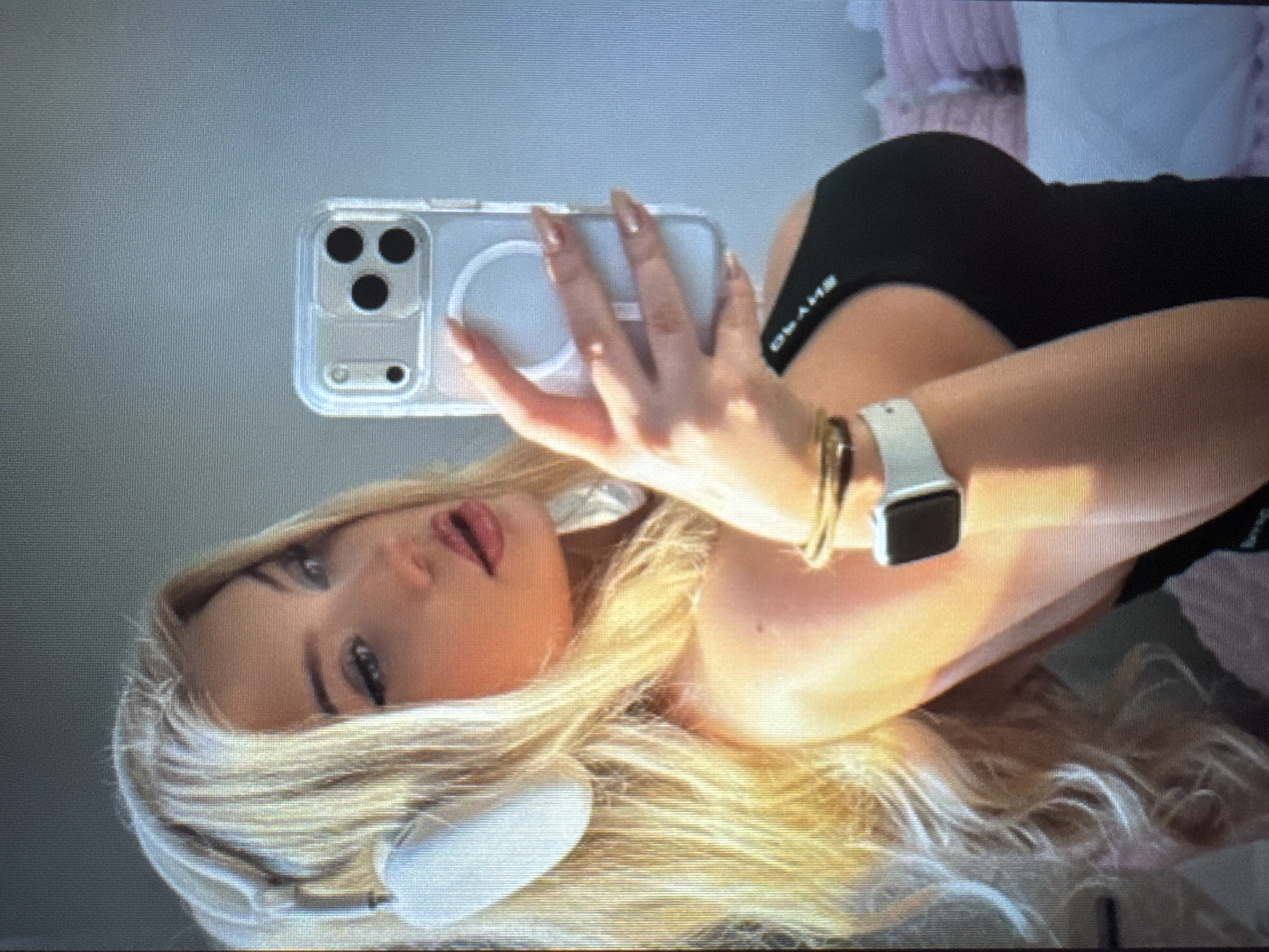 The image, rotated 90 degrees counter-clockwise, depicts a young woman taking a mirror selfie.
**Subjects:** The primary subject is a woman with long, flowing blonde hair, fair skin, and a made-up face featuring dark eye makeup and pink lipstick.
**Objects:** She holds a clear-cased smartphone, resembling an iPhone with a triple camera system, with her right hand, showcasing long, light-colored fingernails. On her left wrist, she wears a white Apple Watch and two thin gold-colored bracelets. White over-ear headphones are resting around her neck. She is wearing a black top or bra. In the background, soft, light-colored, possibly ruffled fabric (like a blanket or pillow) is visible.
**Scene:** The setting appears to be indoors, likely a personal space such as a bedroom, given the soft furnishings in the background. The selfie is taken through a reflection, indicating a mirror or a reflective surface.
**Activity or event taking place:** The woman is actively taking a selfie, holding her phone to capture her reflection in the mirror.
**Notable details:** Her blonde hair is a prominent feature, cascading around her. Her facial expression is a subtle pose, with slightly parted lips. The lighting is bright and even, suggesting it is daytime. The image displays a slight pixelation, possibly indicating it is a photograph of a screen.
**Visible text:** Reversed white text is visible on her black top. When un-reversed, it reads "GLAMB".
