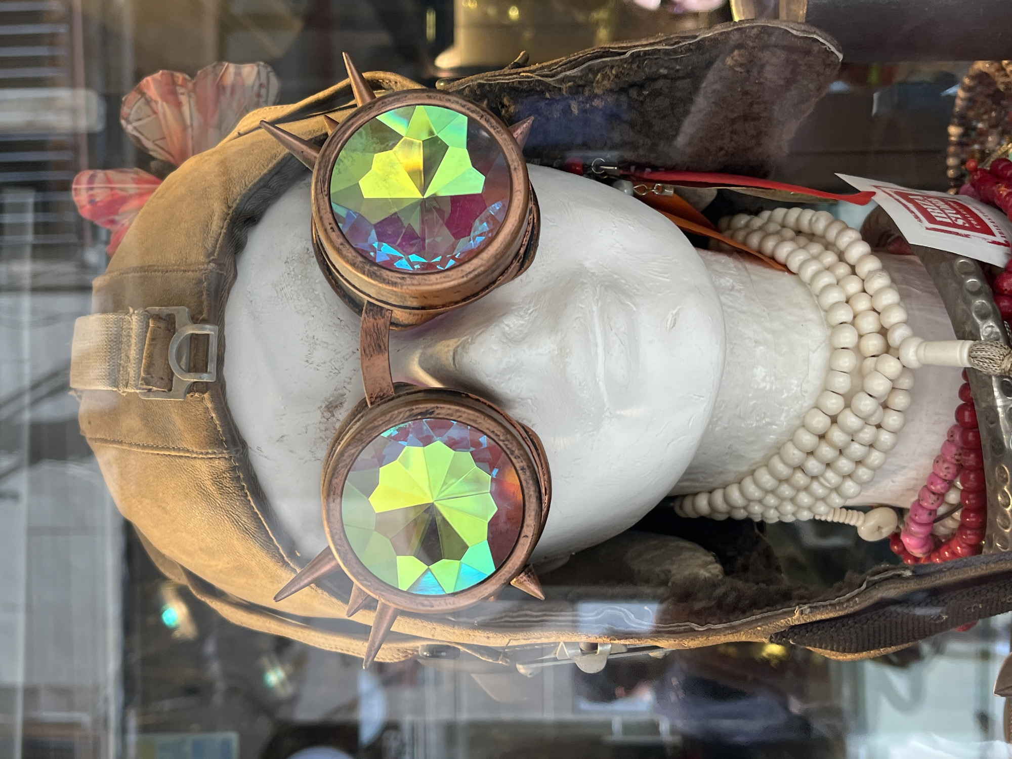 The image displays a static retail or exhibition arrangement, likely within a glass display case or shop window. A white mannequin head, oriented horizontally, is the central subject. It wears a vintage-style aviator pilot's helmet made of worn, light brown leather with a canvas chin strap and a metal buckle.
Dominating the front of the helmet are a striking pair of steampunk-inspired goggles. These goggles feature prominent copper-toned frames, each adorned with small, sharp studs or spikes around the edges. Their large, multifaceted lenses exhibit a vibrant, iridescent quality, refracting light into dazzling patterns of yellow-green, blue, and magenta hues.
To the upper left of the helmet, two decorative elements resembling paper flowers or butterflies, one pink and the other with a subtle pattern, are visible. To the right of the mannequin head, a collection of accessories is clustered. This includes several strands of beads: a thick string of smooth, cream-colored spherical beads, and finer strands of red and pink beads. A silver-colored, textured bracelet or cuff is also present. A small white tag with red text, possibly reading "SUYA", is partially visible among the beads.
The background is softly blurred with reflections, indicating the presence of glass and suggesting a retail environment, such as a shop in Barcelona, Spain, specializing in unique accessories or vintage items. No activity is depicted; it is a still-life arrangement.