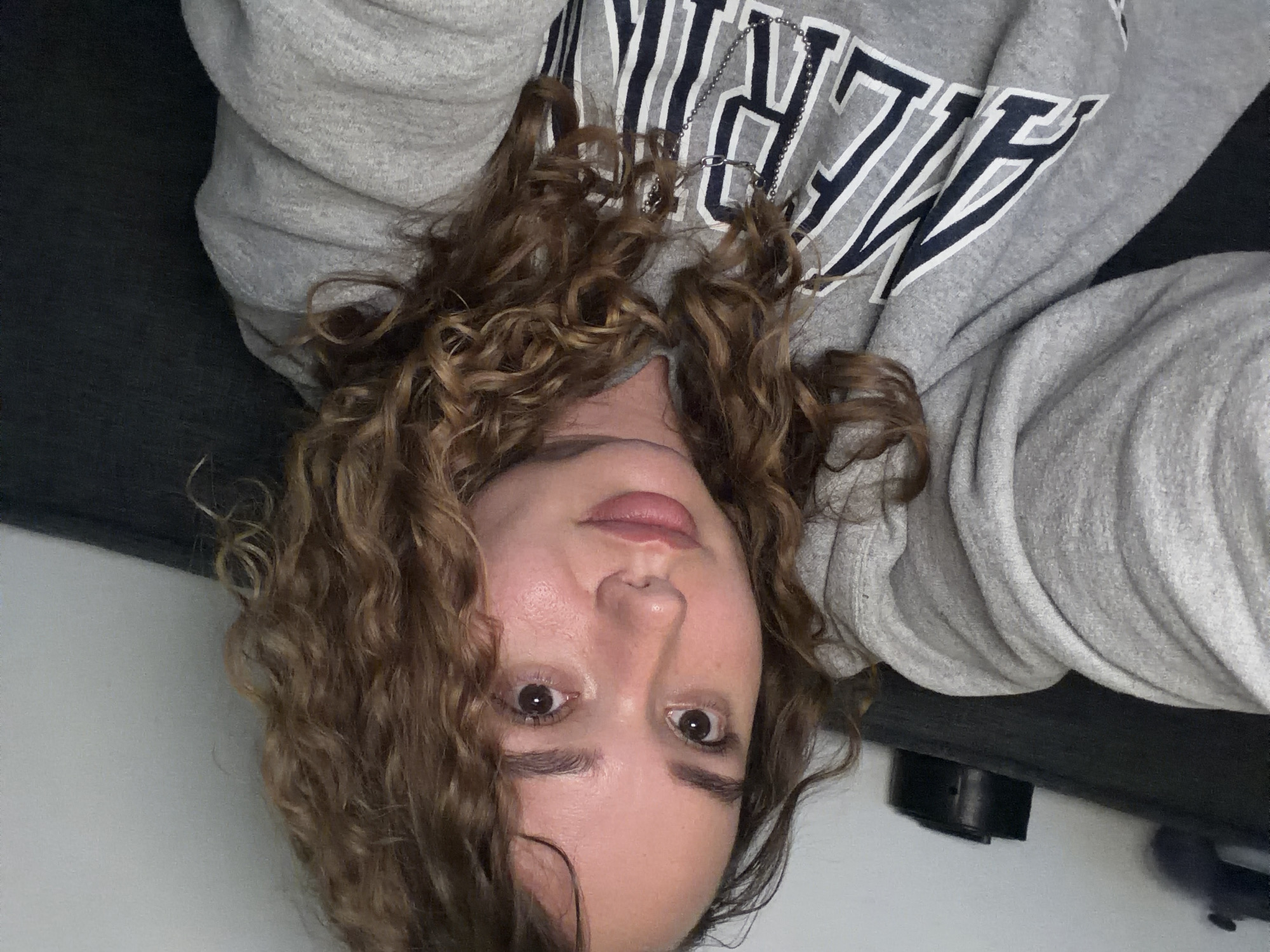 A woman with curly brown hair is lying down, looking up at the camera. She is wearing a grey sweatshirt with navy blue lettering. She is wearing a necklace. Part of a black object is visible. The location is unknown and there is no indication that the image was taken in Ramat Gan, Israel.
