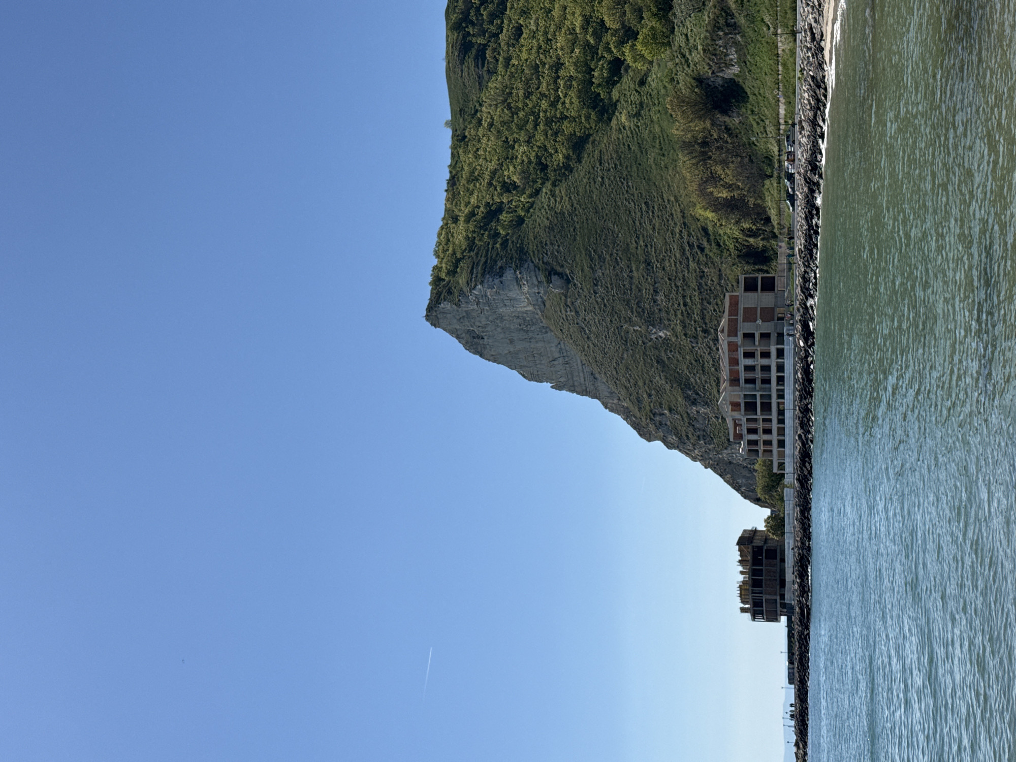 The image shows a coastal scene in Kavarna, Bulgaria. A cliff covered in vegetation slopes down to a shoreline, adjacent to a body of water. There are two unfinished buildings visible along the coast. The sky is clear and blue.