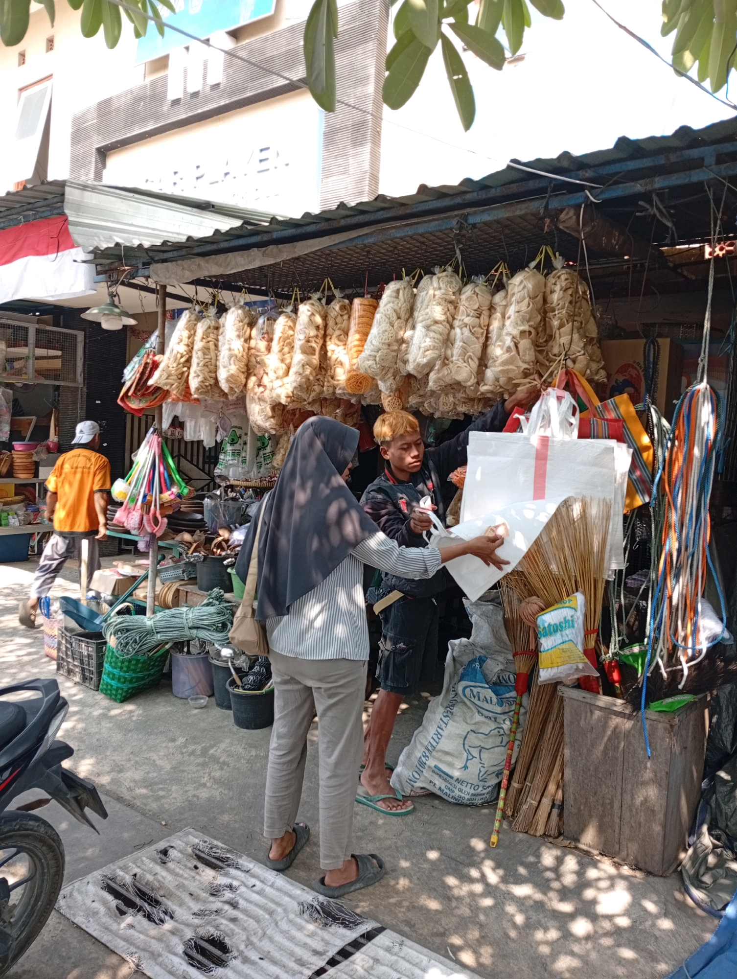 The image shows a street market scene in Playen, Indonesia. In the foreground, a woman wearing a headscarf and a striped shirt is interacting with a young man who has blonde hair and is wearing a dark jacket. They are standing in front of a market stall that is selling various goods. The stall has a corrugated metal roof, and hanging from it are many bags of what appear to be dried snacks or crackers.
Behind the woman, there is a man in an orange t-shirt and grey pants, walking away from the camera. To the left of the stall, shelves are stocked with plastic containers and other items. Various items are displayed on the ground around the stall, including brooms, bags of what looks like animal feed, and coils of rope. In the background, a building with the word "PLAYEN" visible on its facade suggests the location. The ground is paved with concrete, and there are shadows cast by the sun and the market stall. A motorcycle is partially visible on the left side of the image.
