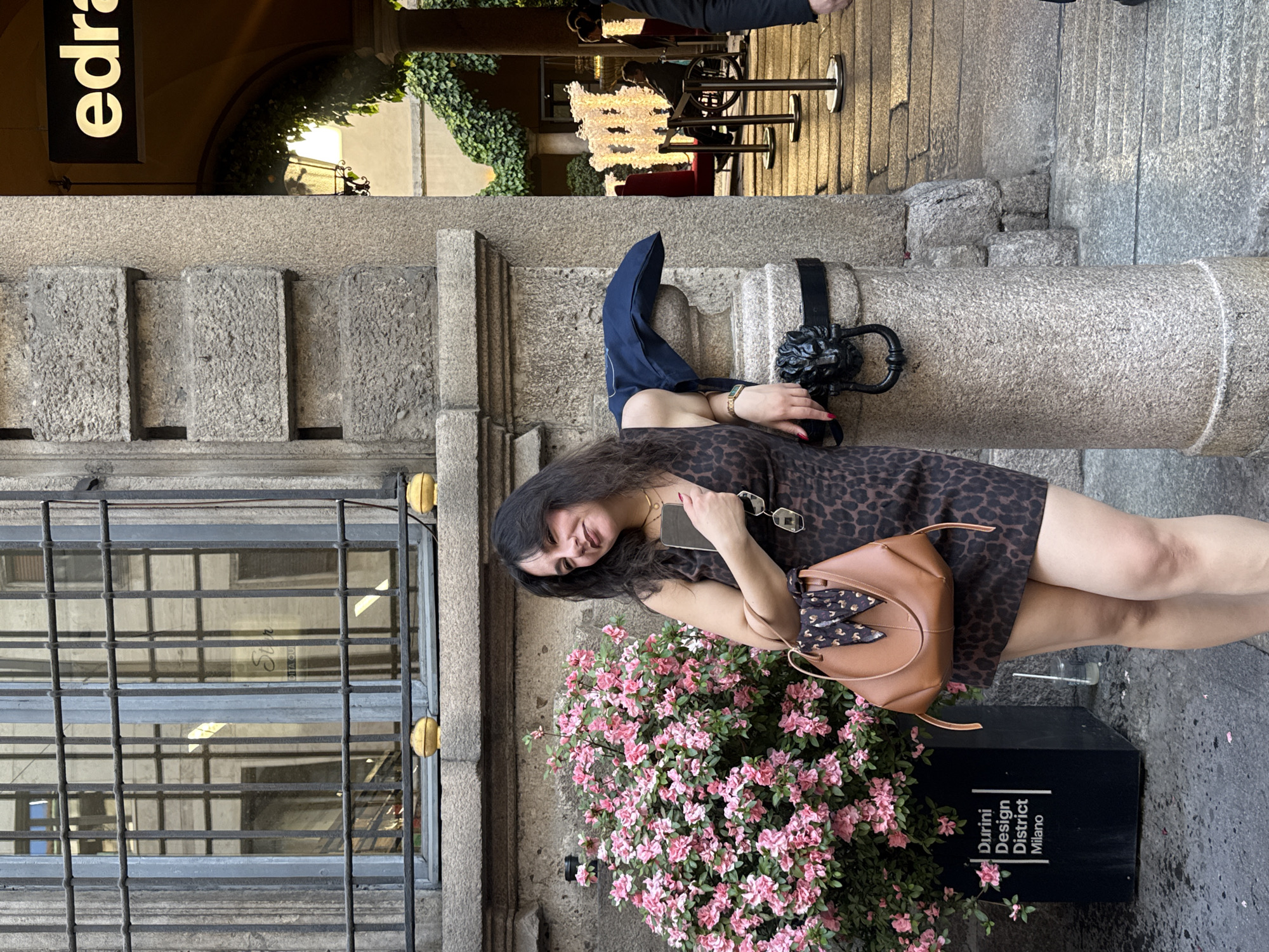 A young woman stands outdoors in an urban setting, posing for the camera with a pleasant, smiling expression. She has dark hair and is wearing a brown and black leopard print short dress. She holds a smartphone in her right hand and sunglasses in her left, which rests on a light brown structured handbag adorned with a patterned scarf. Gold jewelry, including a necklace and bracelet, and red nail polish are also visible.
She stands next to a large black planter filled with vibrant pink azalea-like flowers. White text on the planter reads "Durini Design District Milano", indicating the location in Milan, Italy. Behind her to the right is a rough-hewn stone pillar with a decorative black metal fixture. To her left, a window covered by a dark metal grate with yellow accents is visible.
In the background, an archway draped with green ivy leads to an outdoor cafe or restaurant area. Tables, chairs, and a distinctive illuminated white, cloud-like art installation or light fixture can be seen. Above the archway, a black sign with white lettering displays "edra". The overall lighting suggests it is daytime, possibly late afternoon, and the weather appears clear. The scene combines traditional stone architecture with modern urban elements.