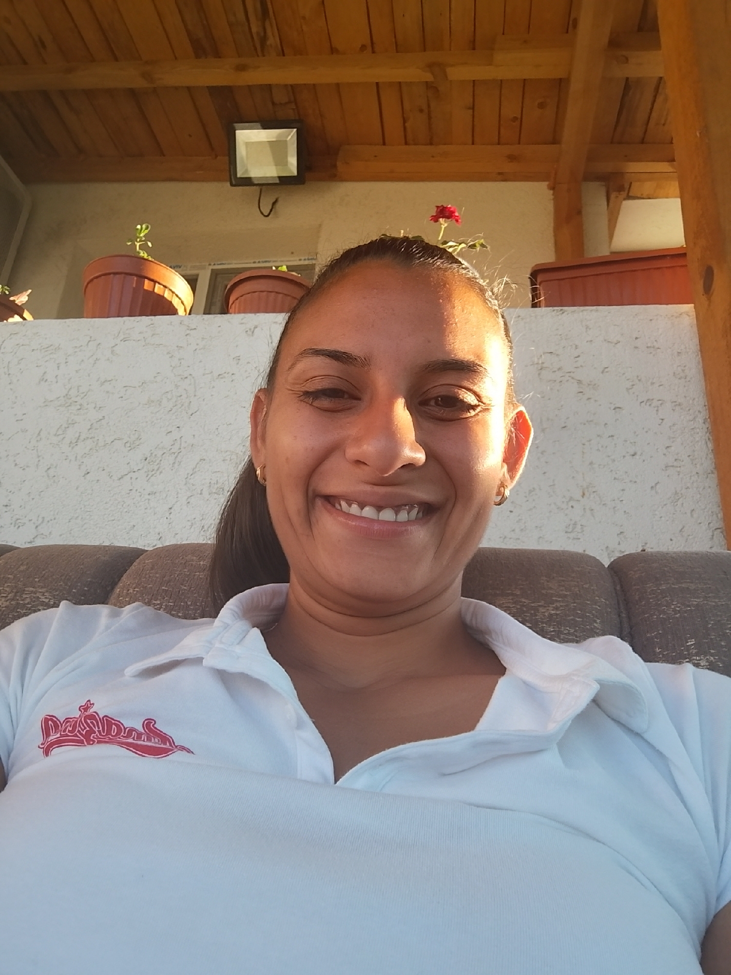 A woman with dark hair pulled back and a gold hoop earring in her visible left ear smiles broadly at the camera, her head tilted slightly. She wears a white polo shirt with a red logo on the left side of the chest. She is seated on a brown upholstered couch, with the back of the couch visible behind her. The background features a textured white wall, several terra cotta pots containing plants, and a wooden-beamed overhang. A black outdoor light fixture is mounted on the wall. The lighting suggests it is daytime, with sunlight casting shadows. The scene is outdoors, likely a patio or porch area, and the woman appears relaxed and happy.