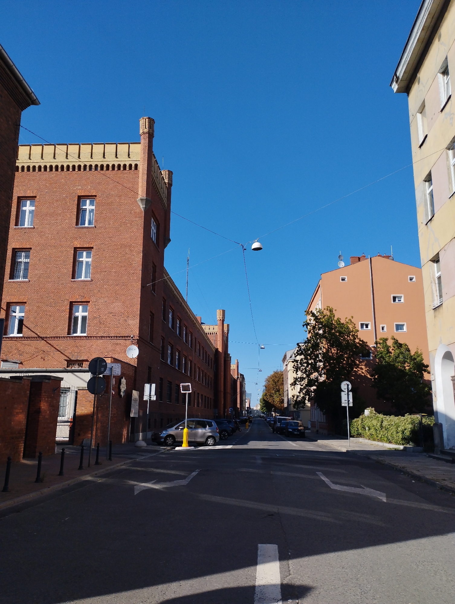 The image shows a street scene in Szczecin, Poland, under a clear blue sky. On the left is a tall, brick building with multiple windows and decorative crenellations at the top. This building appears to be an older structure, possibly with a historical or institutional function. Adjacent to it, also on the left, is a smaller brick wall and a gateway.
To the right of the street, across from the brick building, is a multi-story building painted in a warm, orangey-brown color. This building also has numerous windows of varying sizes. Further to the right, another building with a pale yellow facade is visible, featuring arched doorways at its base.
The street itself is paved with asphalt and marked with white directional arrows indicating traffic flow. Several cars are parked along the sides of the street and on the road. Traffic lights and street lamps are visible, suspended over the road by wires. Trees with dense foliage are present, casting shadows on the street and the buildings. The overall impression is of an urban environment on a bright, sunny day.