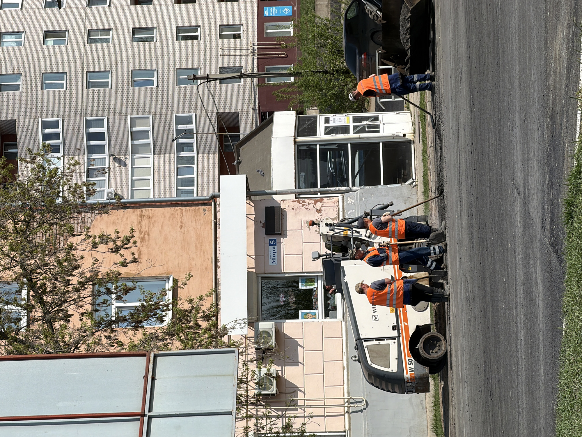 The image depicts a street maintenance scene in Votkinsk, Russia. Three workers wearing orange vests and dark trousers are working on a newly paved section of road. They are using tools and equipment, including a white and black vehicle labeled "W 50." The workers appear to be spreading or smoothing asphalt.
Behind the workers, there is a building with a light pink facade. The building has multiple windows, some of which have posters visible inside. A sign on the building reads "Mira 5" in Russian. Air conditioning units are attached to the building's exterior.
In the background, a tall grey building with numerous windows is visible. The sky is bright, suggesting it is daytime. To the right of the road is a strip of green grass. Part of a parked black car is seen in the upper-right corner.