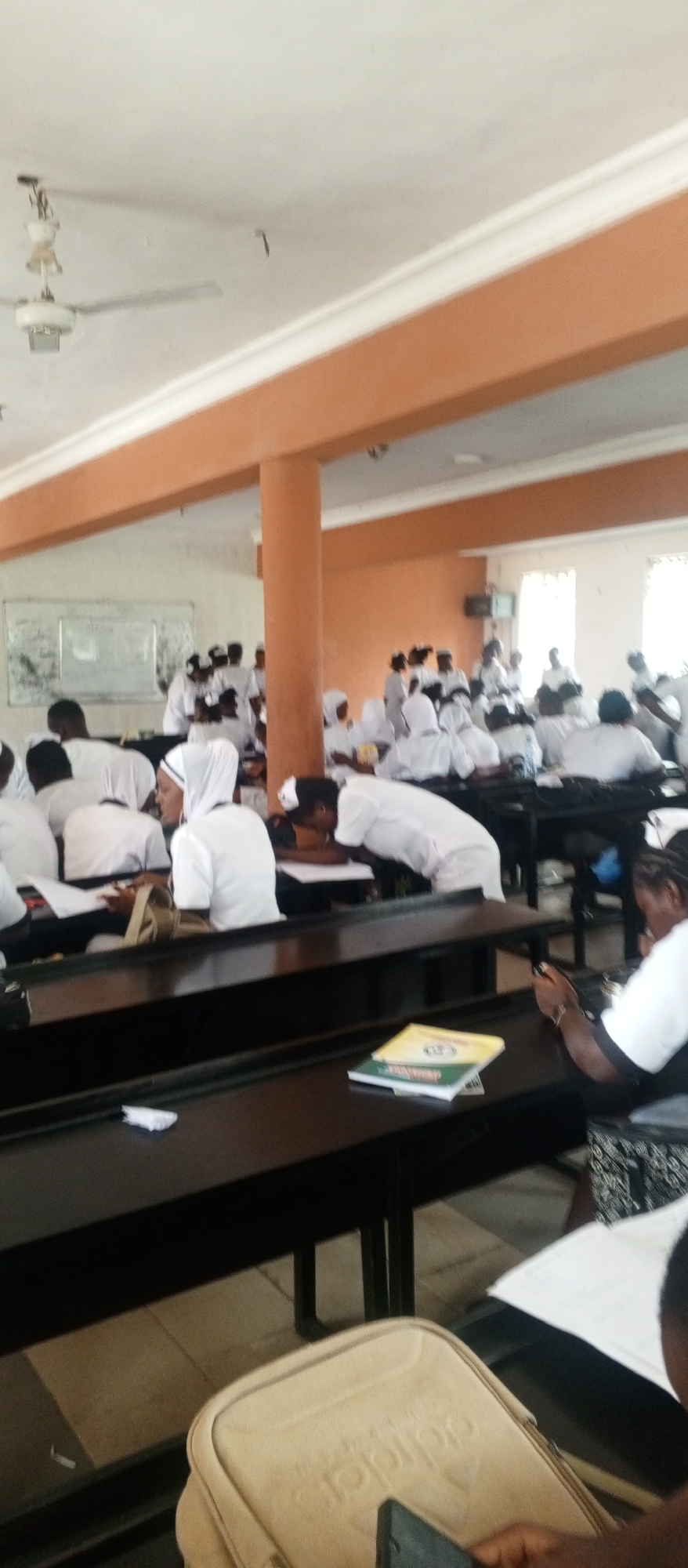 This media file depicts a classroom scene with a group of people, likely students, attending a lecture or studying. The setting appears to be a large room with rows of dark wooden desks and benches. A white ceiling fan is visible, suggesting an indoor environment. The walls are painted in a light color, with a band of orange trim running along the top. In the background, a whiteboard is mounted on the wall, though the writing on it is illegible.
The subjects are predominantly young adults, many of whom are wearing white attire, suggesting a uniform or specific event such as a religious gathering or a medical/nursing class. Some individuals are seated at desks, while others are standing or interacting. Several people are looking down, possibly at notes or their phones. One person in the foreground is holding a smartphone and a light-colored bag with a visible embossed logo that appears to be "adidas". Another person is leaning over a desk, appearing to be writing or studying.
The lighting in the room suggests it is daytime, possibly late morning or afternoon, with natural light entering from windows that are not clearly visible. The overall mood seems to be one of focus and academic activity. The presence of books on the desks further reinforces the educational context. There are no obvious external environmental cues like weather. The lack of clear identifying features makes it difficult to pinpoint an exact location.