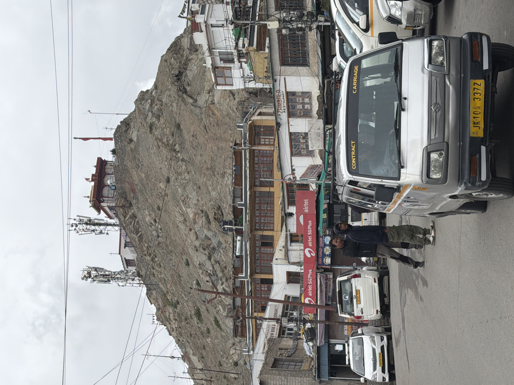 The image depicts a street scene in Leh, India, with a distinctive rocky hillside dominating the background. Atop the hill, several structures are visible, including buildings with red roofs and what appear to be telecommunications towers. The buildings on the hillside are a mix of traditional and modern architecture, with prayer flags also present.
In the foreground, the street is lined with various vehicles, including a silver Maruti Suzuki van with the text "CONTRACT CARRIAGE" on its windshield, two white SUVs, and other cars. Two men are standing on the street near the vehicles, one wearing a dark jacket and the other a grey tracksuit. Behind them, shops and buildings with signs such as "airtel 5GPlus" and "EAT IN ZAMBALA RESTAURANT" indicate commercial activity. The sky is overcast, suggesting a cloudy or partly cloudy day. The overall impression is a bustling urban environment set against a dramatic natural landscape.