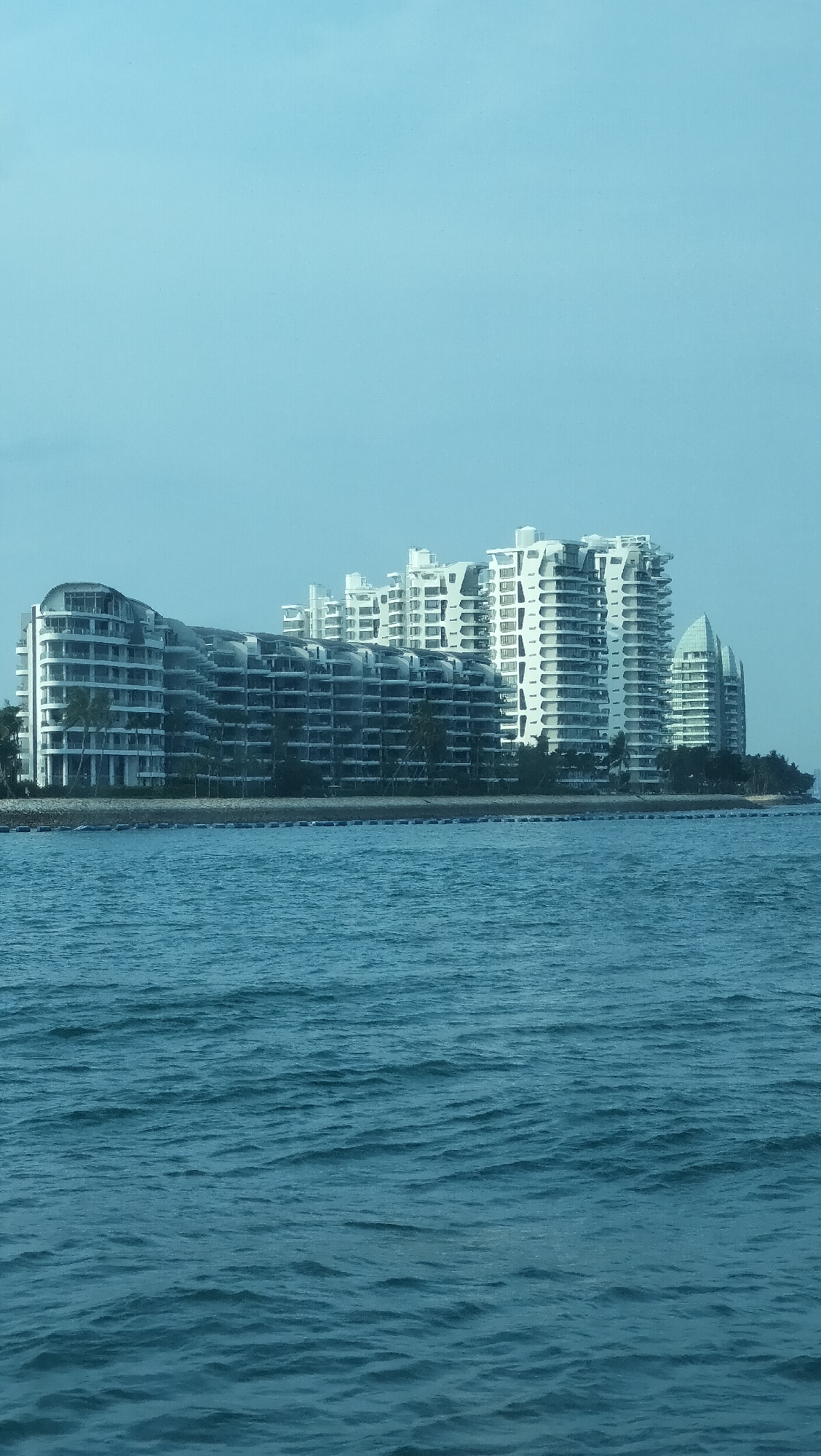 The image shows a panoramic view of the coastal skyline of Singapore, featuring a cluster of modern, white residential buildings. The buildings have a distinct architectural style with curved facades and multiple balconies, suggesting a luxury condominium complex. They are situated along a shoreline, with a line of buoys visible in the water between the buildings and the camera. The sea in the foreground is a deep blue with visible waves, indicating a partly cloudy or hazy day. The buildings appear to be designed with an emphasis on natural light and open spaces, with many windows and balconies. The overall impression is one of contemporary urban development integrated with a coastal environment.
