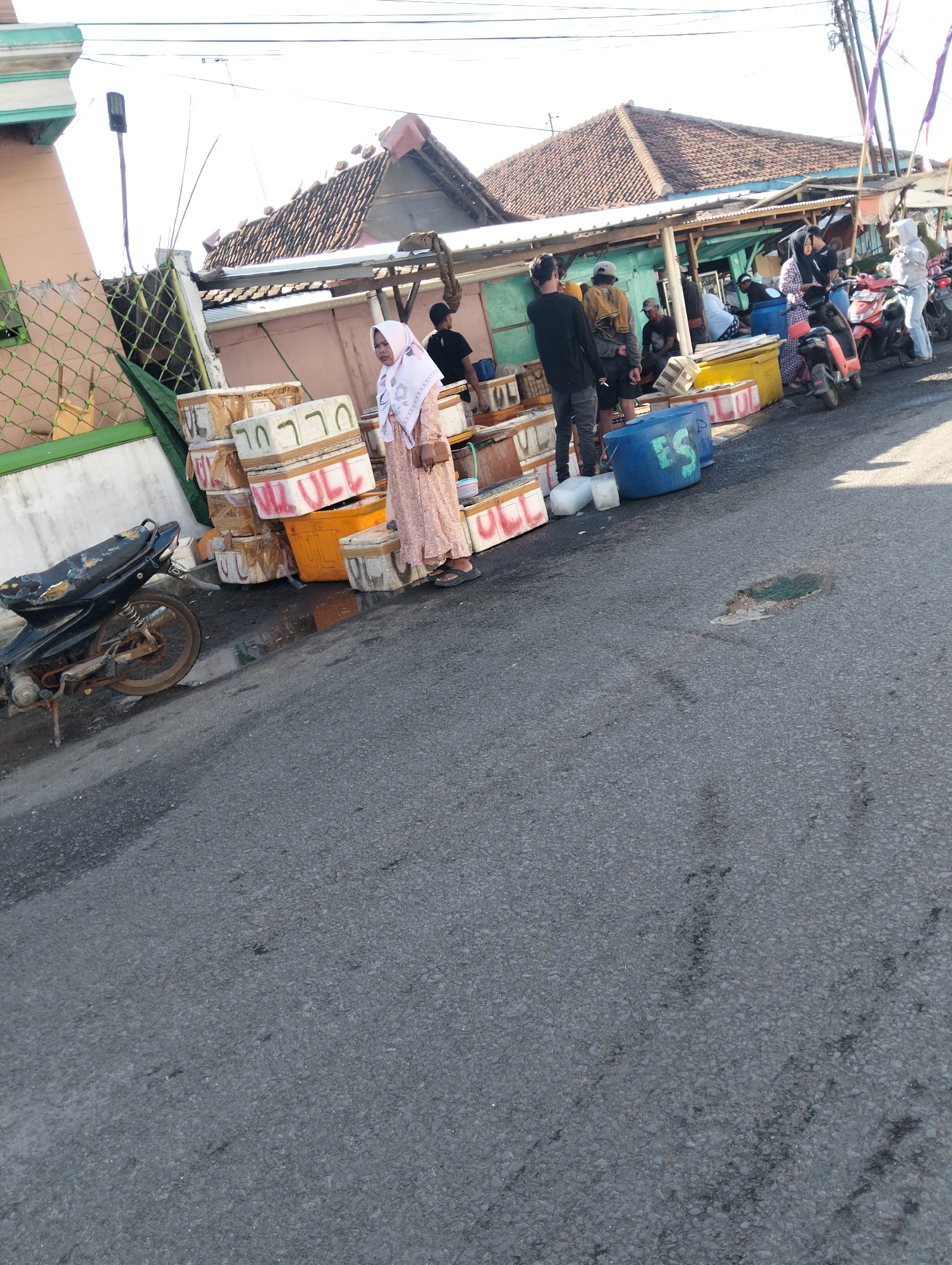 The image shows a street scene in Muncar, Indonesia, with a focus on a fish market or distribution point. In the foreground, a weathered black motorcycle is parked on the asphalt road, its kickstand down. To the right of the motorcycle, a woman in a light pink patterned dress and a headscarf stands next to stacks of white and orange plastic containers, some with red writing, suggesting they are for transporting goods, likely fish.
Behind the woman, several people are visible. Men in casual clothing are standing and interacting around more stacked containers and blue plastic drums. The background reveals a row of market stalls or shops with corrugated metal roofs. The architecture of the buildings suggests a tropical environment. Sunlight casts shadows, indicating it is daytime. The overall scene depicts a busy outdoor commercial area, characteristic of a coastal town where fishing is a primary activity.