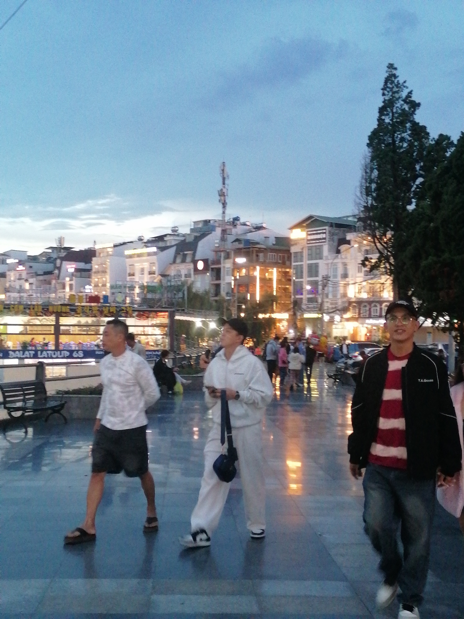 The image depicts an outdoor scene in Da Lat, Vietnam, at dusk or twilight. The ground is paved, appearing wet or reflective, and illuminated by scattered lights from nearby buildings. Three men are the primary subjects in the foreground:
1. **Man 1 (left):** Middle-aged, wearing a white long-sleeved shirt and black shorts, walking with his hands behind his back. He is wearing sandals.
2. **Man 2 (center):** Wearing a white tracksuit and carrying a small bag over his shoulder. He has short dark hair and appears to be looking towards the right.
3. **Man 3 (right):** Wearing a black jacket over a red and white striped shirt, blue jeans, and a black baseball cap. He is wearing glasses.
In the background, there are several buildings with various lights on, indicating commercial or residential use. The architecture suggests a hillside town. Some buildings have multi-story structures and appear to be hotels or businesses.
Several other people are visible in the background, walking, standing, or sitting, suggesting a public gathering place. There are some trees to the right, as well as benches. The atmosphere is generally serene, with soft, diffused lighting.