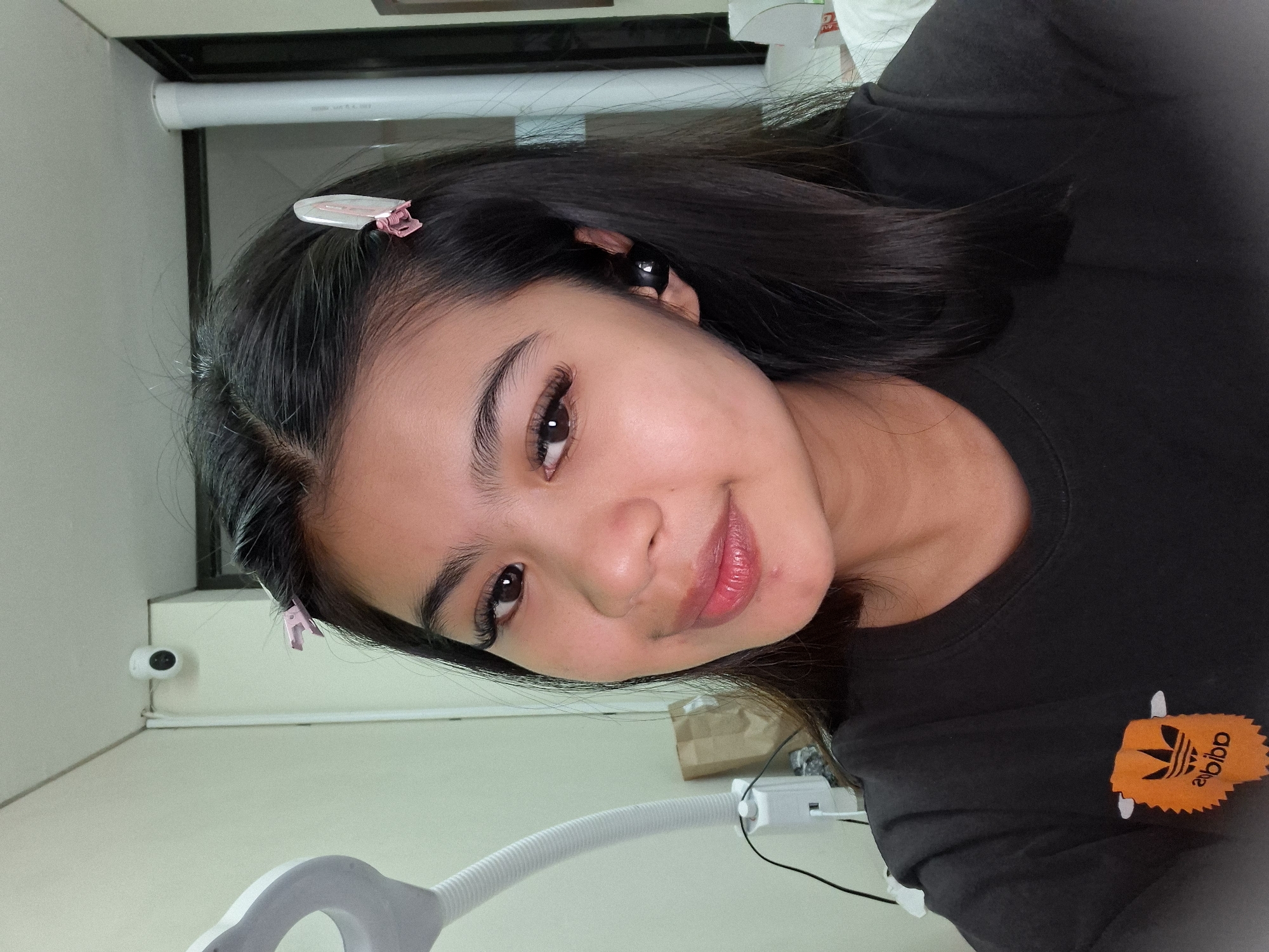 A young woman with long, dark hair is taking a selfie in what appears to be an indoor studio or treatment room. She is wearing a black t-shirt with an orange Adidas logo. Pink hair clips are visible in her hair, and she has on black wireless earbuds. A security camera is mounted on the wall behind her, and a white lamp is to her left. The background includes a wall, a doorway, and a white pillar. The image does not provide any specific information about the location in Kuta, Indonesia.